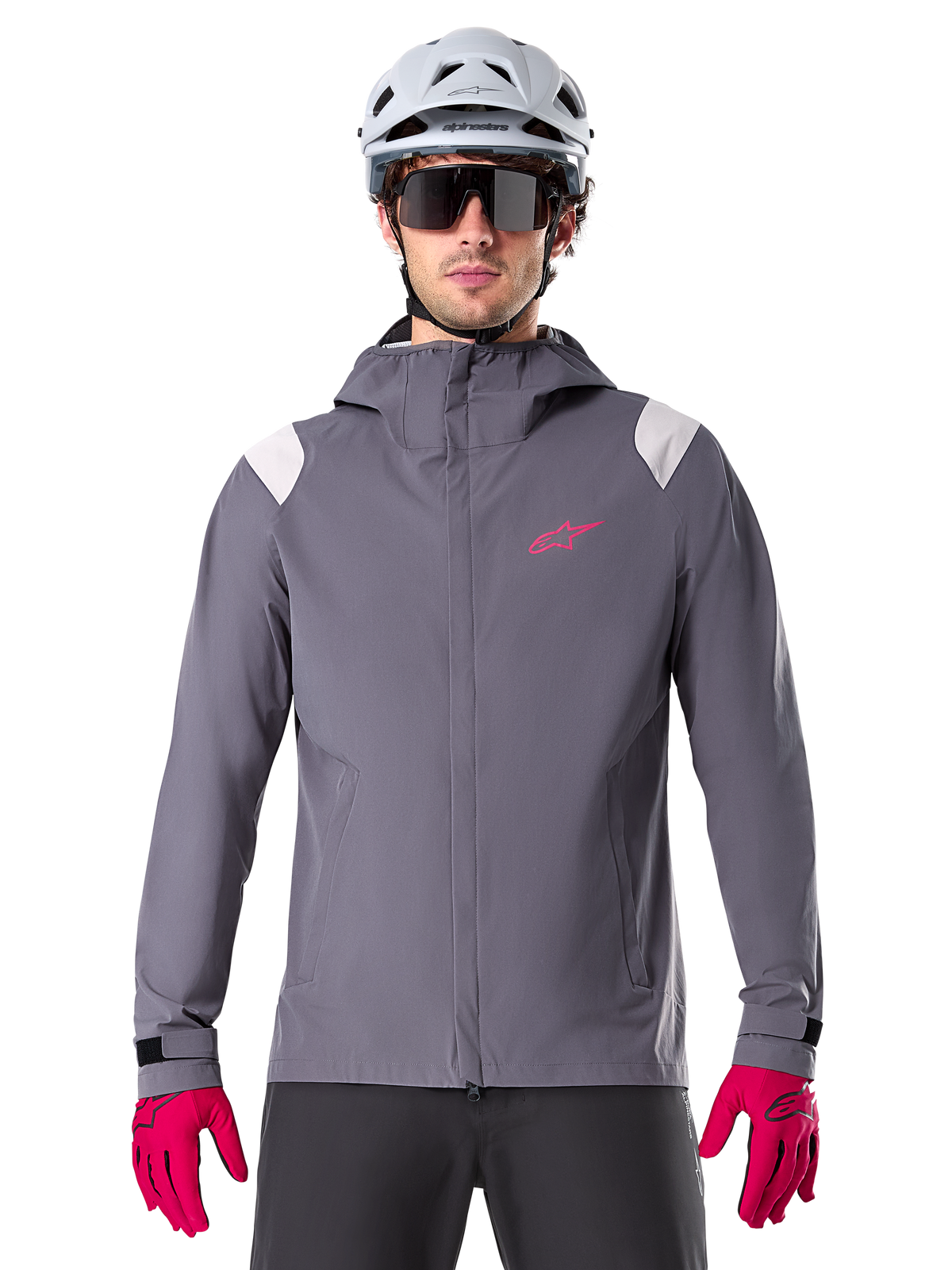 Kurtka przeciwdeszczowa Alpinestars A-Dura, rowerowa kurtka przeciwdeszczowa MTB, kolor ciemny tytanowy szary, fason z kapturem z różowym logo Alpinestars i jasnoszarymi panelami na ramionach, noszona z białym kaskiem MTB i jaskraworóżowymi rękawiczkami, wodoodporna odzież do jazdy w terenie