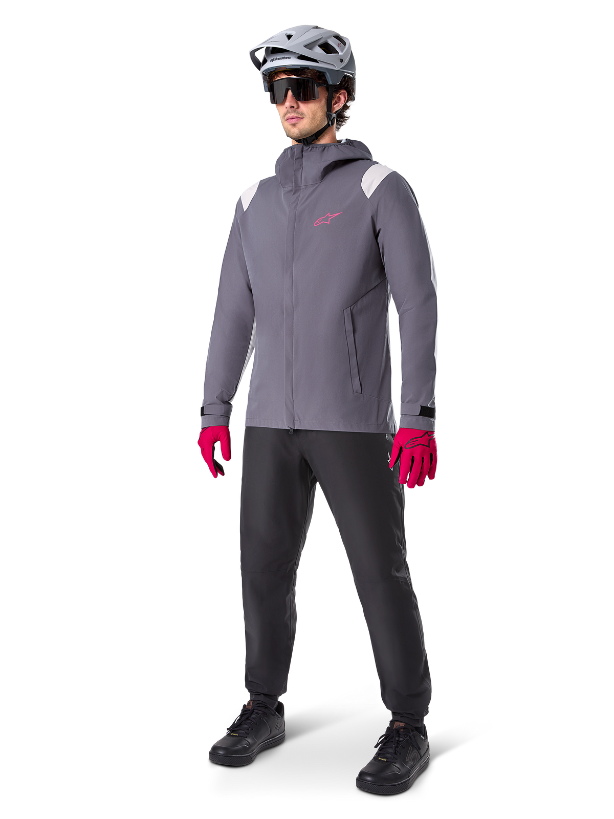 Model w kurtce przeciwdeszczowej Alpinestars A-Dura w kolorze Dark Titanium, szara kurtka z kapturem z różowym logo na piersi i jasnoszarymi panelami na ramionach, zestawiona z szarym kaskiem MTB, ciemnymi okularami, jaskraworóżowymi rękawicami i czarnymi spodniami do jazdy terenowej.