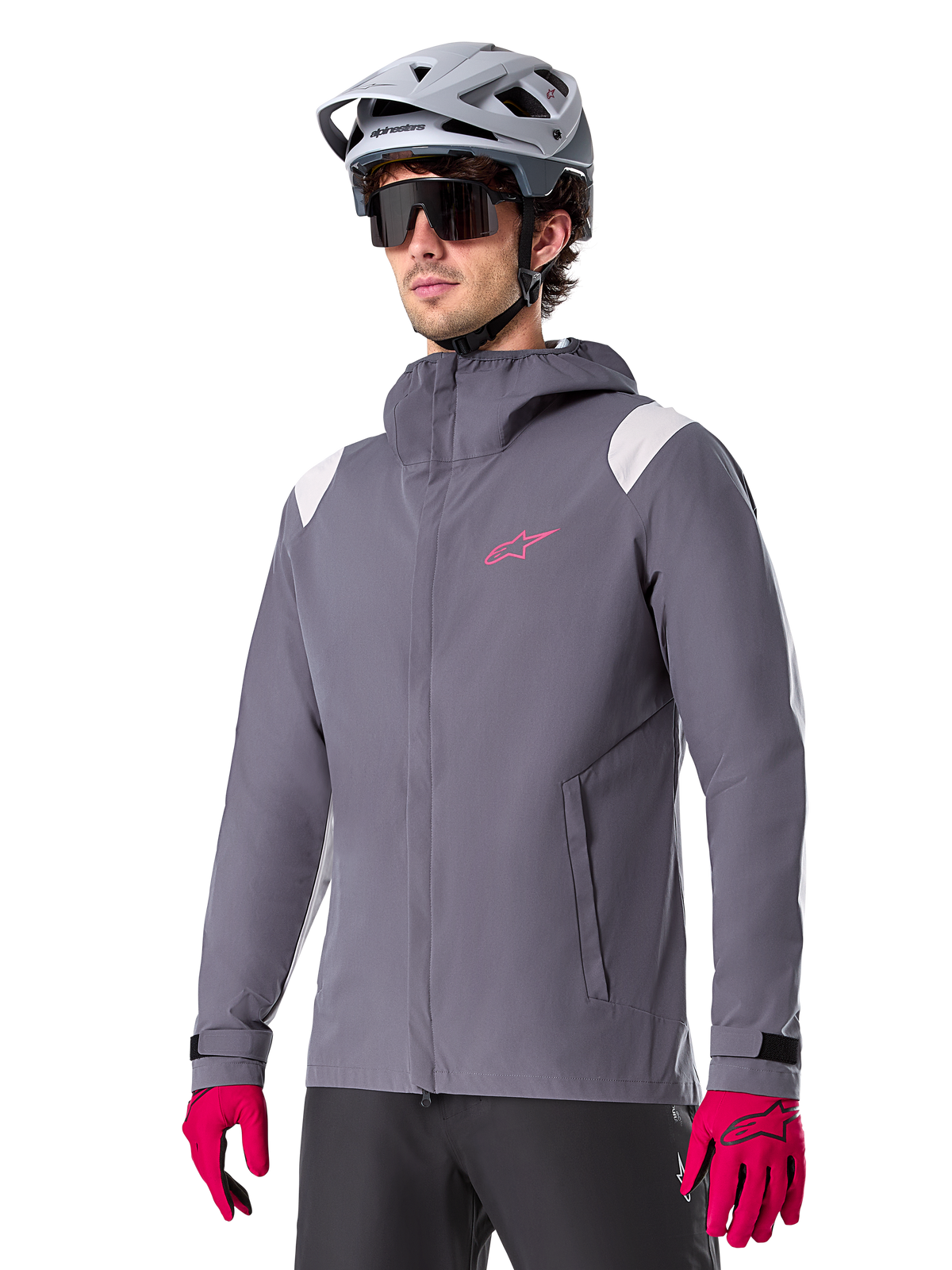 Alpinestars A-Dura Rain Jacket, rowerowa kurtka przeciwdeszczowa MTB, ciemnoszara z różowym logo i białymi akcentami na ramionach, model z kapturem i przednimi kieszeniami, noszona z różowymi rękawiczkami i jasnoszarym kaskiem rowerowym