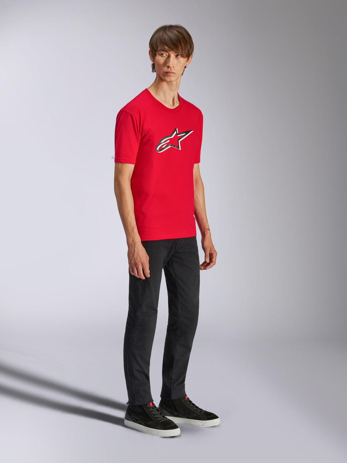 Alpinestars Ageless Shadow CSF Tee, czerwona koszulka z krótkim rękawem, z dużym logo Ageless na piersi, noszona przez modela w czarnych spodniach i sneakersach, codzienna odzież streetwear w stylu motorsport