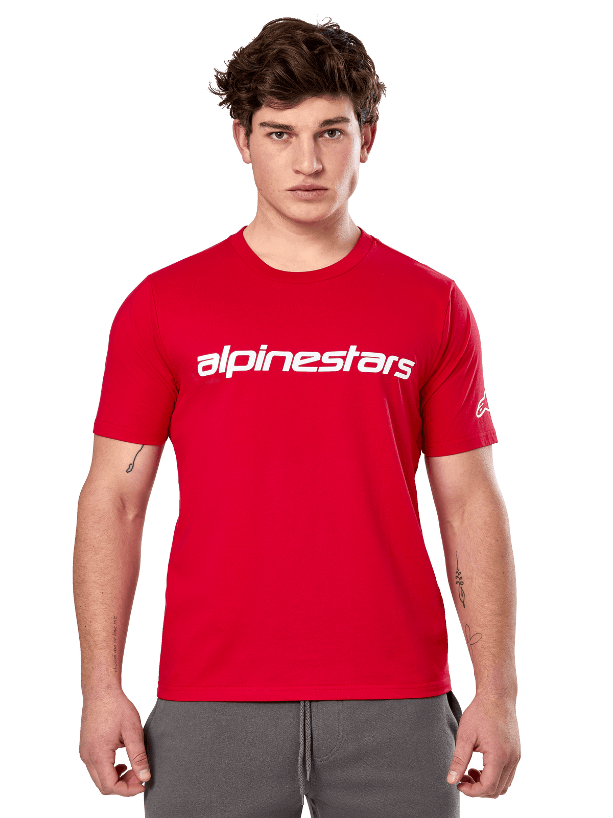 Alpinestars Linear Wordmark 2.0 CSF Tee, czerwona koszulka z krótkim rękawem, biały linearny znak słowny na klatce piersiowej i logo Ageless na rękawie, model w szarych spodniach dresowych, casualowy styl streetwear inspirowany motorsportem