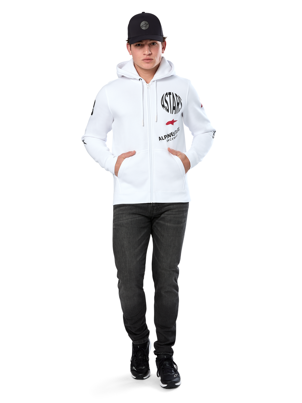 Bluza z kapturem Alpinestars Variegate, biała rozpinana bluza polarowa z kapturem, wieloelementowa grafika z napisem „ASTARS” i czerwonym logo gwiazdy na piersi, detale graficzne na rękawach, model w czarnej czapce z daszkiem i czarnych jeansach, codzienna odzież streetwear w stylu motorsport