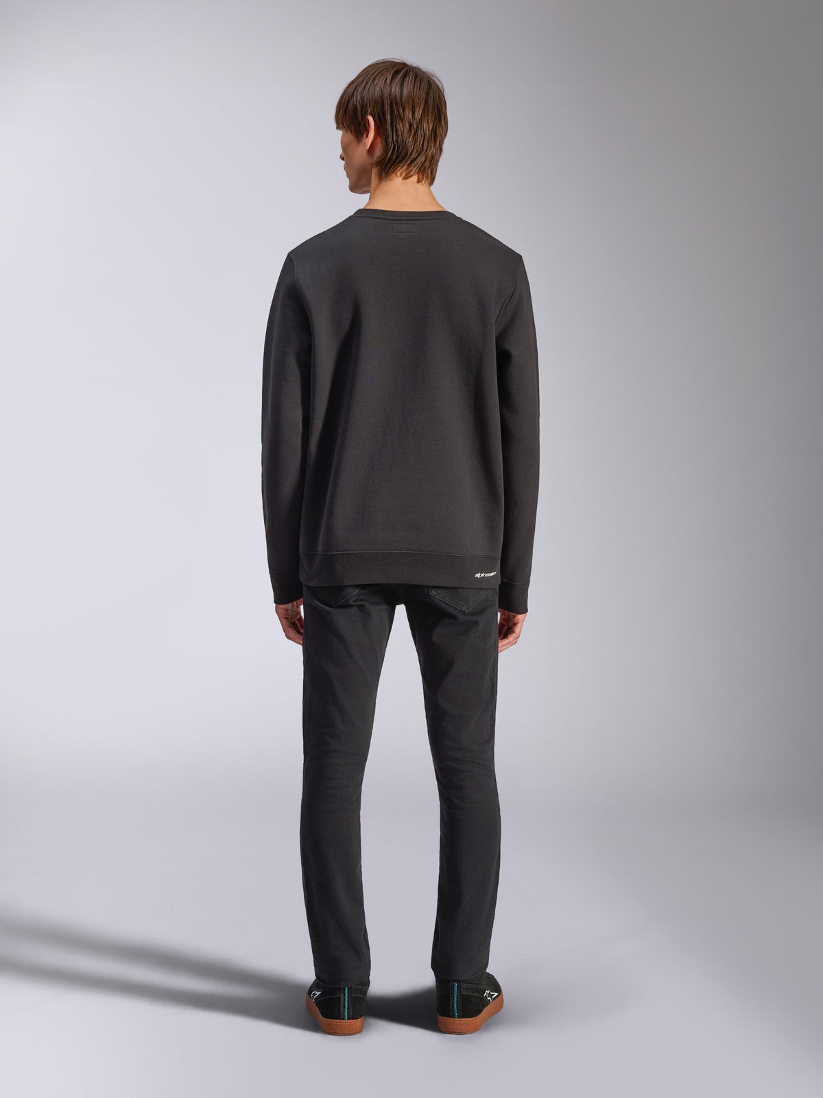 Alpinestars Positive Crew, czarna bluza typu pullover, widok z tyłu na męskim modelu, zestawiona z czarnymi jeansami i casualowymi sneakersami, wykończona ściągaczami przy mankietach i na dole, streetwear inspirowany motorsportem