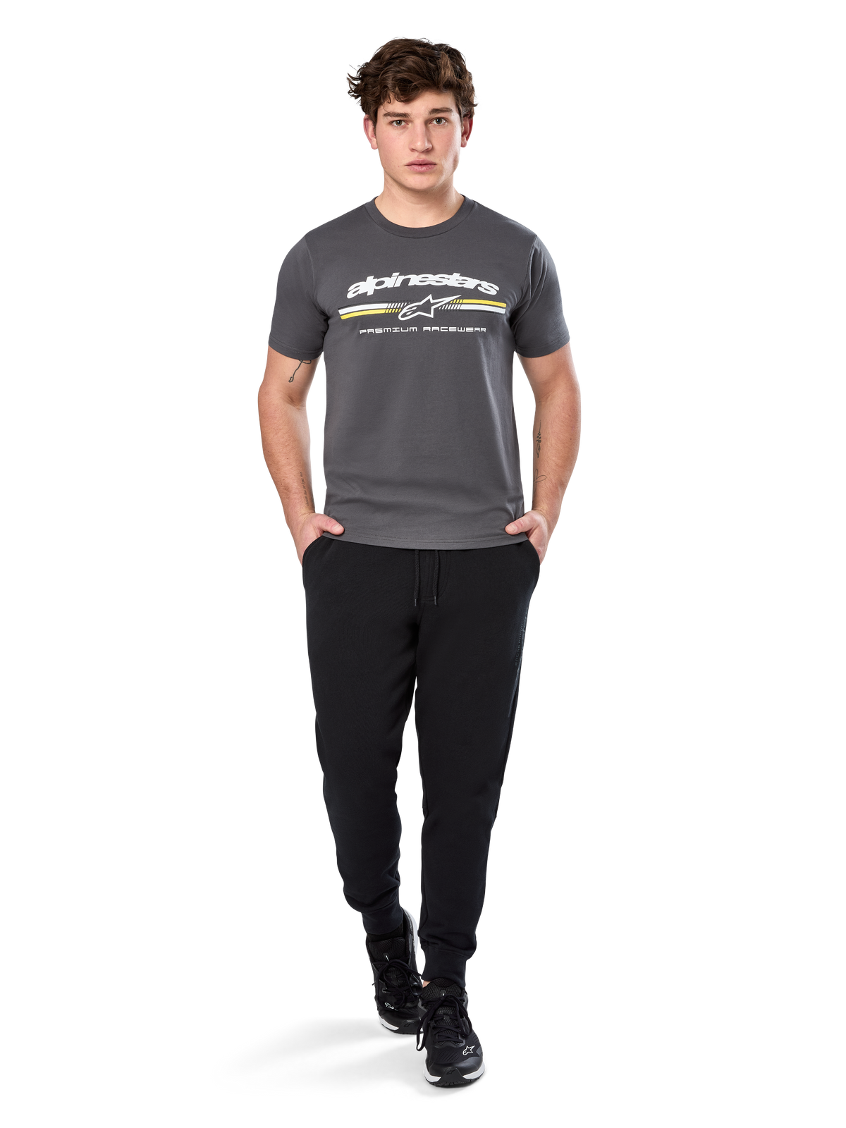 Mężczyzna w koszulce Alpinestars Prevail Csf Tee z krótkim rękawem, kolor grafitowy, z białym logo Alpinestars i trójkolorowym paskiem z żółtymi akcentami, w zestawieniu z czarnymi joggersami i sneakersami, casualowy streetwear w stylu motorsport