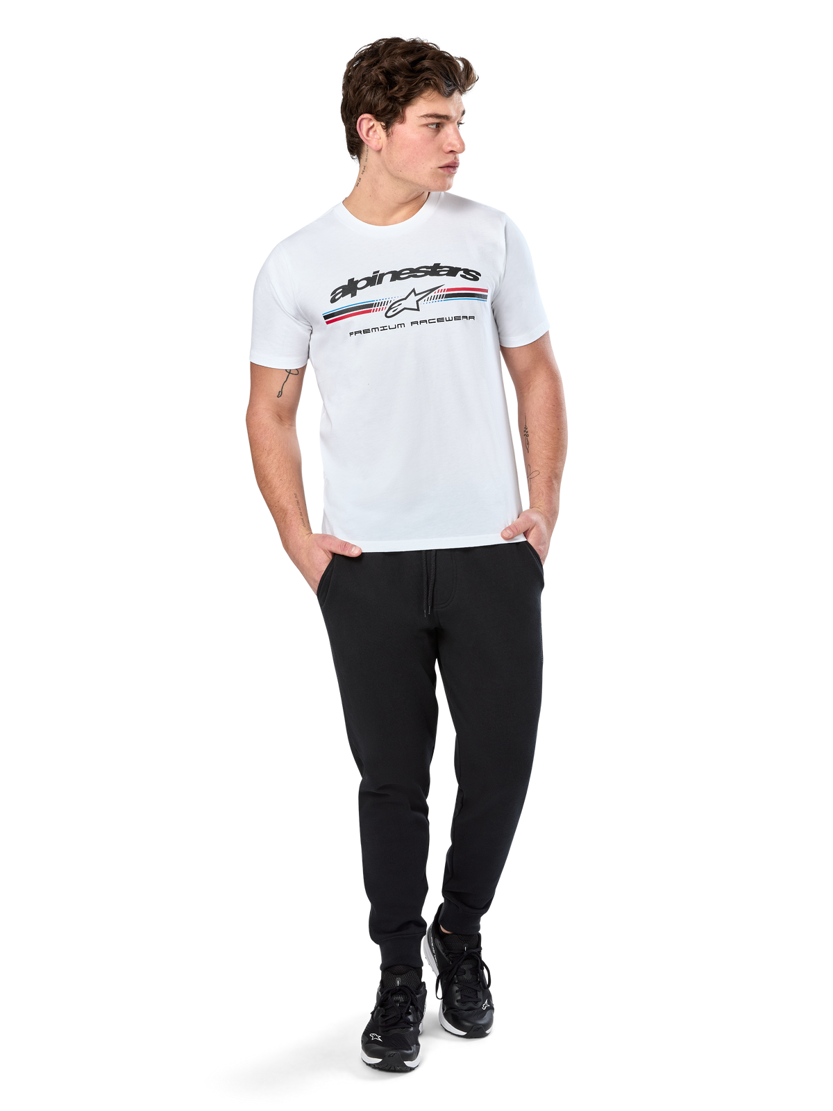 Alpinestars Prevail CSF Tee, koszulka z krótkim rękawem, biała, z logo z trzema kolorowymi paskami w stylu paddock i brandingiem Alpinestars z przodu, prezentowana na modelu w czarnych joggerach i czarnych sneakersach, codzienna moda uliczna w stylu motorsport