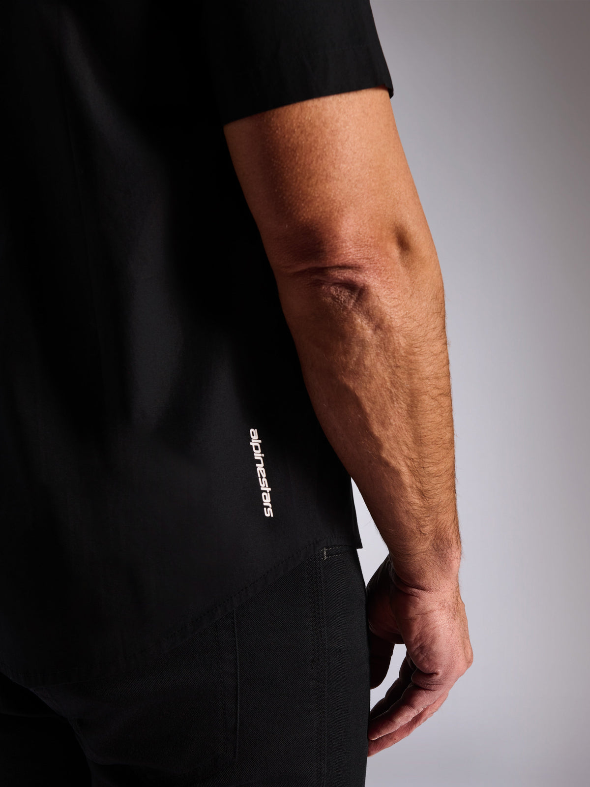 Alpinestars Strive Woven Shirt, czarna koszula z krótkim rękawem zapinana na guziki, pionowe białe logo Alpinestars z boku, zbliżenie na dolną krawędź i logo marki, codzienna odzież uliczna w stylu motorsport