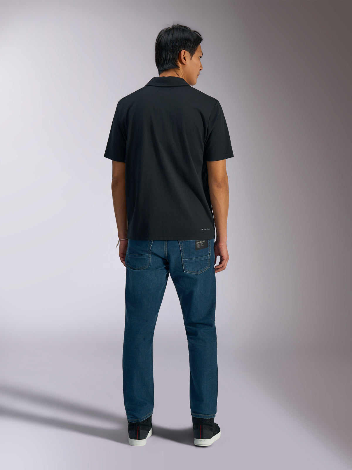 Alpinestars Impetus Sport Polo, Black, back view of short-sleeve polo shirt with Alpinestars branding on the lower back, worn with blue jeans, casual motorsport-inspired sportswear