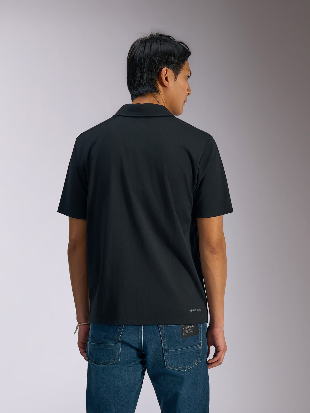 Alpinestars Impetus Sport Polo, Black Short Sleeve Polo Shirt, back view of man wearing polo with blue denim jeans, featuring small Alpinestars wordmark on the lower back, casual motorsport-inspired sportswear