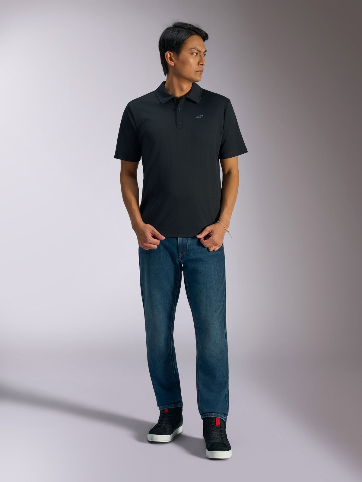 Alpinestars Impetus Sport Polo, Black short sleeve polo shirt, features a three-button placket and grey Astars chest logo, worn by a man with blue jeans and black high-top sneakers, casual motorsport-inspired sportswear