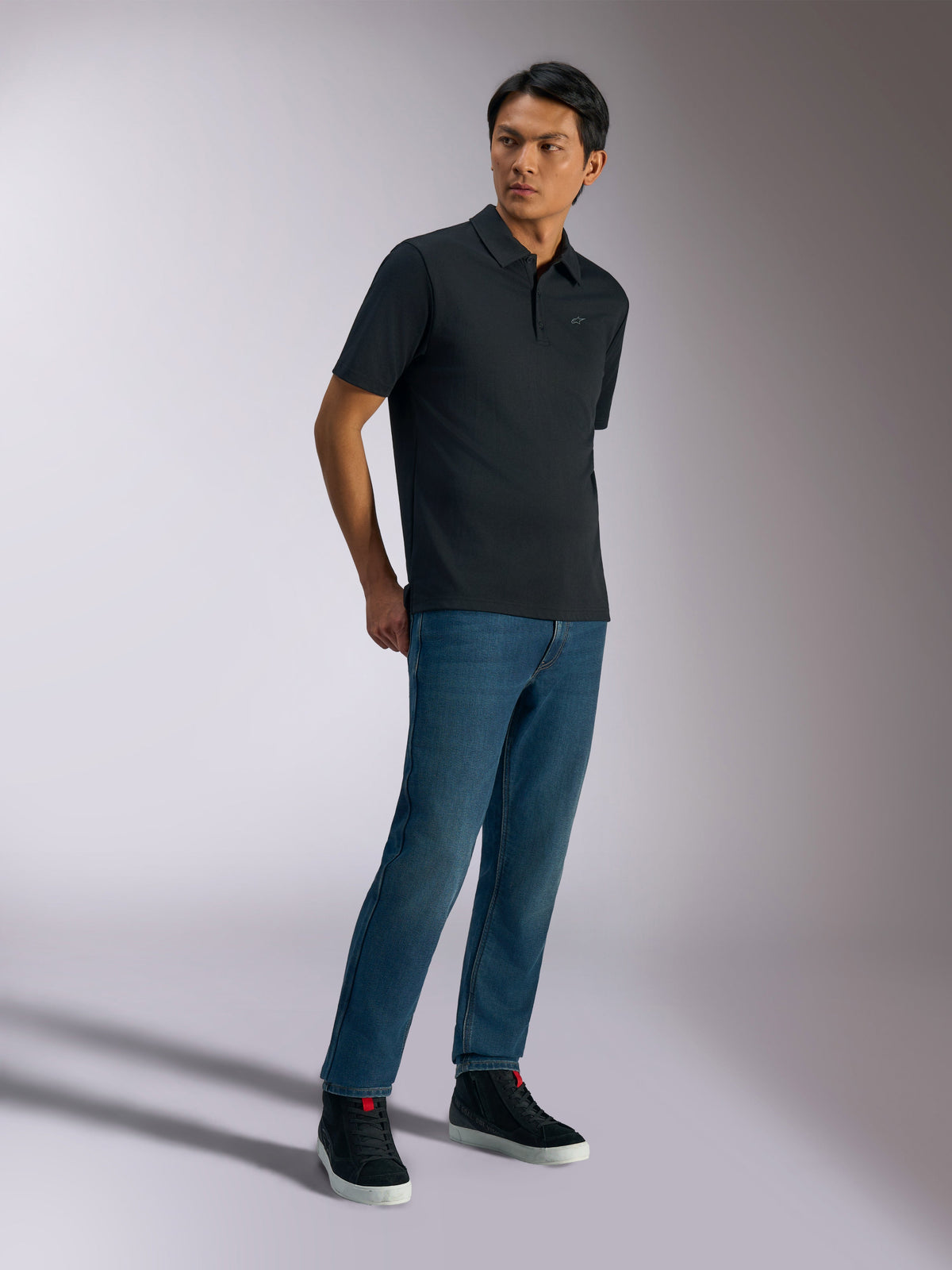 Alpinestars Impetus Sport Polo, Short Sleeve Polo Shirt, Black, featuring a subtle chest logo and three-button placket, worn by a model with blue jeans and black sneakers, casual motorsport-inspired lifestyle apparel