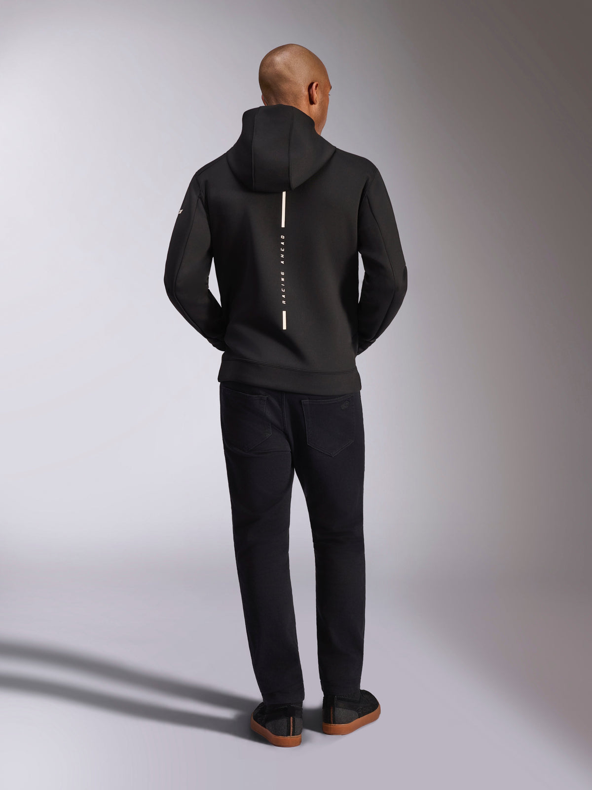 Alpinestars Insight Hoodie, Black, rear view on model, featuring a vertical white back stripe logo with "Racing Ahead" text, casual sportswear hoodie in tri-blend double face fleece, paired with black pants.