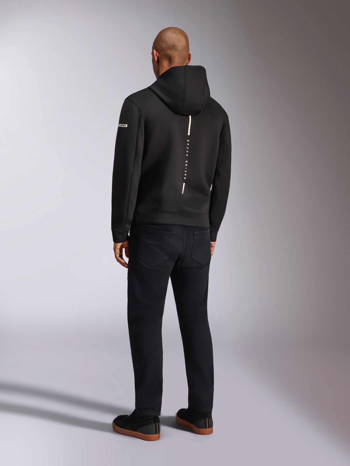 Alpinestars Insight Hoodie, Black sportswear hoodie, back view showing a vertical white stripe with 'RACING AHEAD' branding, sleeve logo, paired with black pants and sneakers, casual motorsport-inspired design
