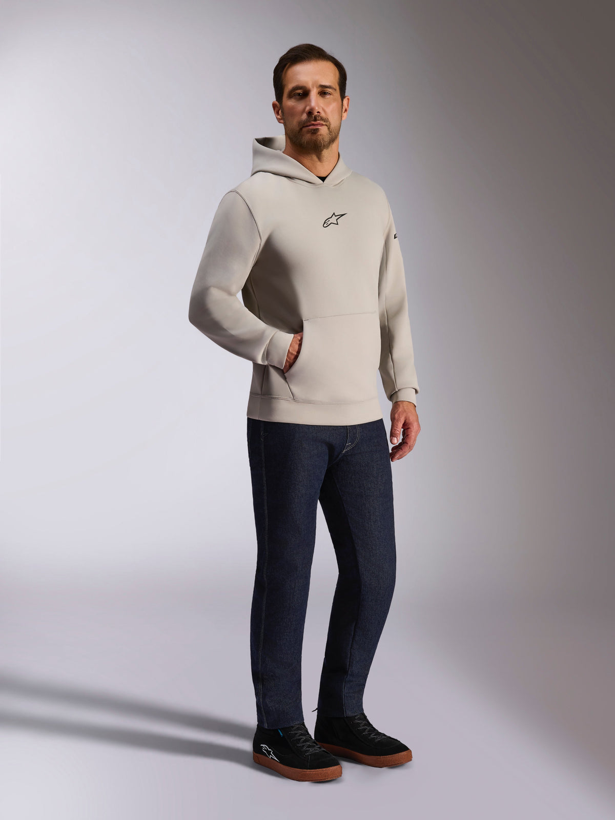Bluza z kapturem Alpinestars Insight, codzienna odzież uliczna, kolor szary cementowy z czarnym logo Astars na piersi, fason wkładany przez głowę z kieszenią kangurką, zestawiona z ciemnoniebieskimi jeansami i czarnymi sneakersami, nowoczesny styl inspirowany sportami motorowymi