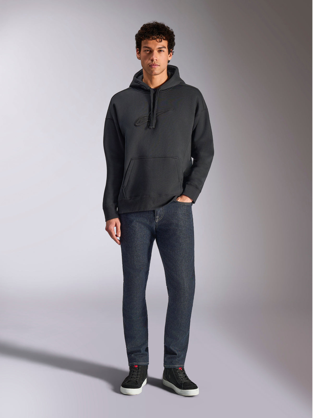 Alpinestars Emergent Oversized Hoodie, casual lifestyle streetwear, black with tonal 3D embroidered logo, drawstring hood, kangaroo pocket, oversized fit, worn by a model with dark blue jeans and black sneakers.