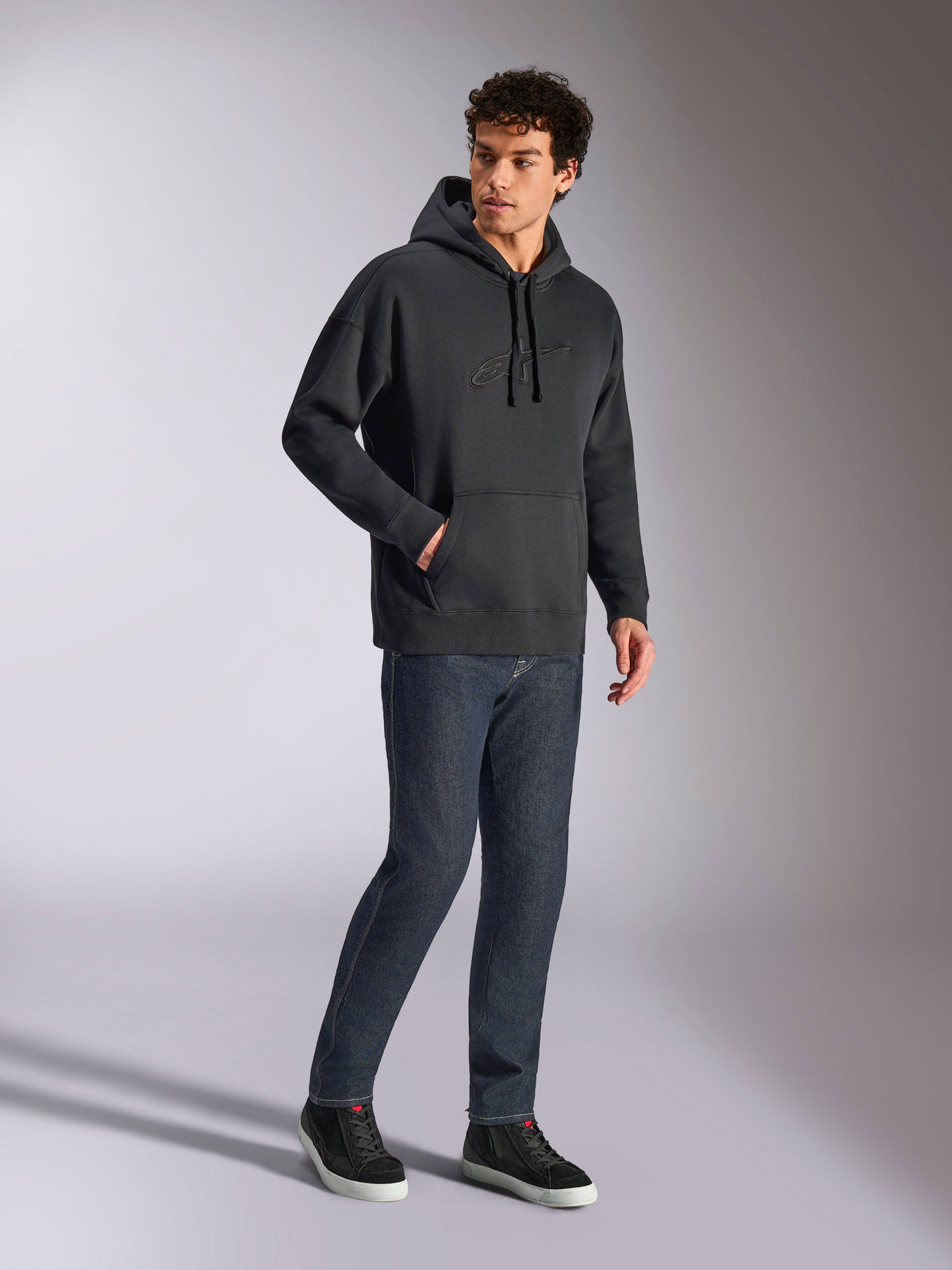 Alpinestars Emergent Oversized Hoodie, black casual streetwear, tonal 3D embroidered Astars logo on chest, kangaroo pocket, oversized fit, worn by male model with dark blue denim jeans and black sneakers