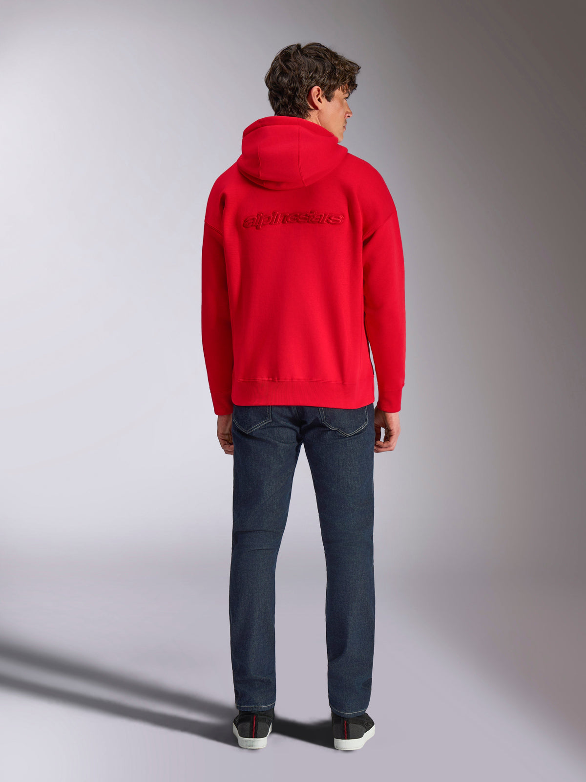 Alpinestars Emergent Oversized Hoodie, czerwona bluza z kapturem, widok z tyłu na modelu, haftowane logo 3D Alpinestars w tym samym kolorze, ciemnoniebieskie dżinsy, codzienna odzież uliczna inspirowana motorsportem