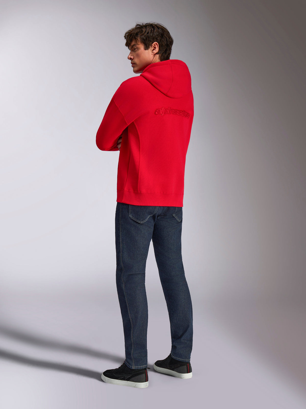 Alpinestars Emergent Oversized Hoodie, czerwona bluza z kapturem, widok z tyłu ukazujący haftowane logo z napisem Alpinestars na plecach, luźny krój, zestawiona z ciemnoniebieskimi dżinsami i czarnymi sneakersami, codzienna stylizacja streetwear