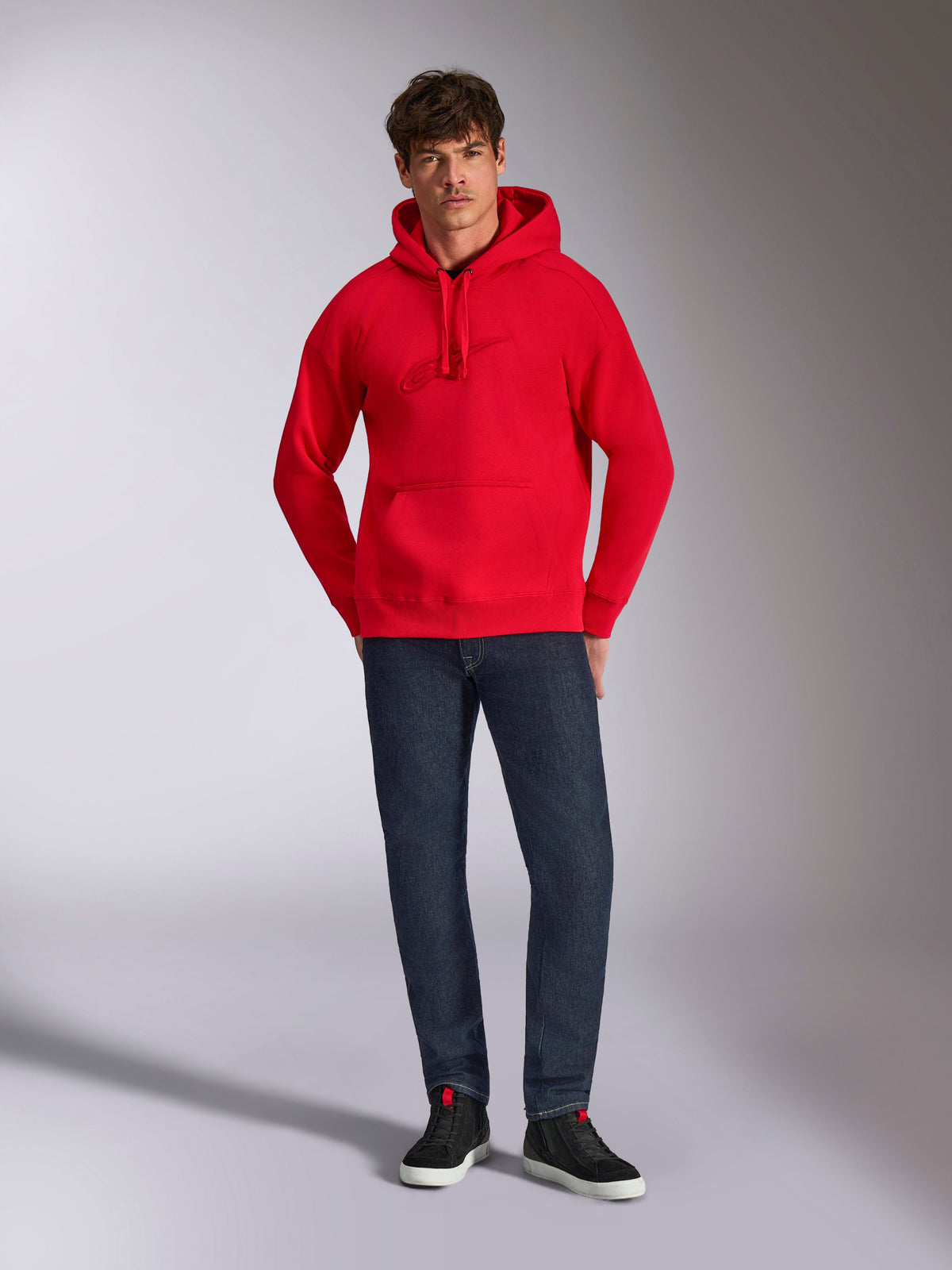 Alpinestars Emergent Oversized Hoodie, czerwona, z tonalnym haftowanym logo Astars 3D na piersi, kieszenią typu kangurka i kapturem ściąganym sznurkiem, noszona przez modela z ciemnoniebieskimi dżinsami i czarnymi trampkami, casualowy styl streetwear