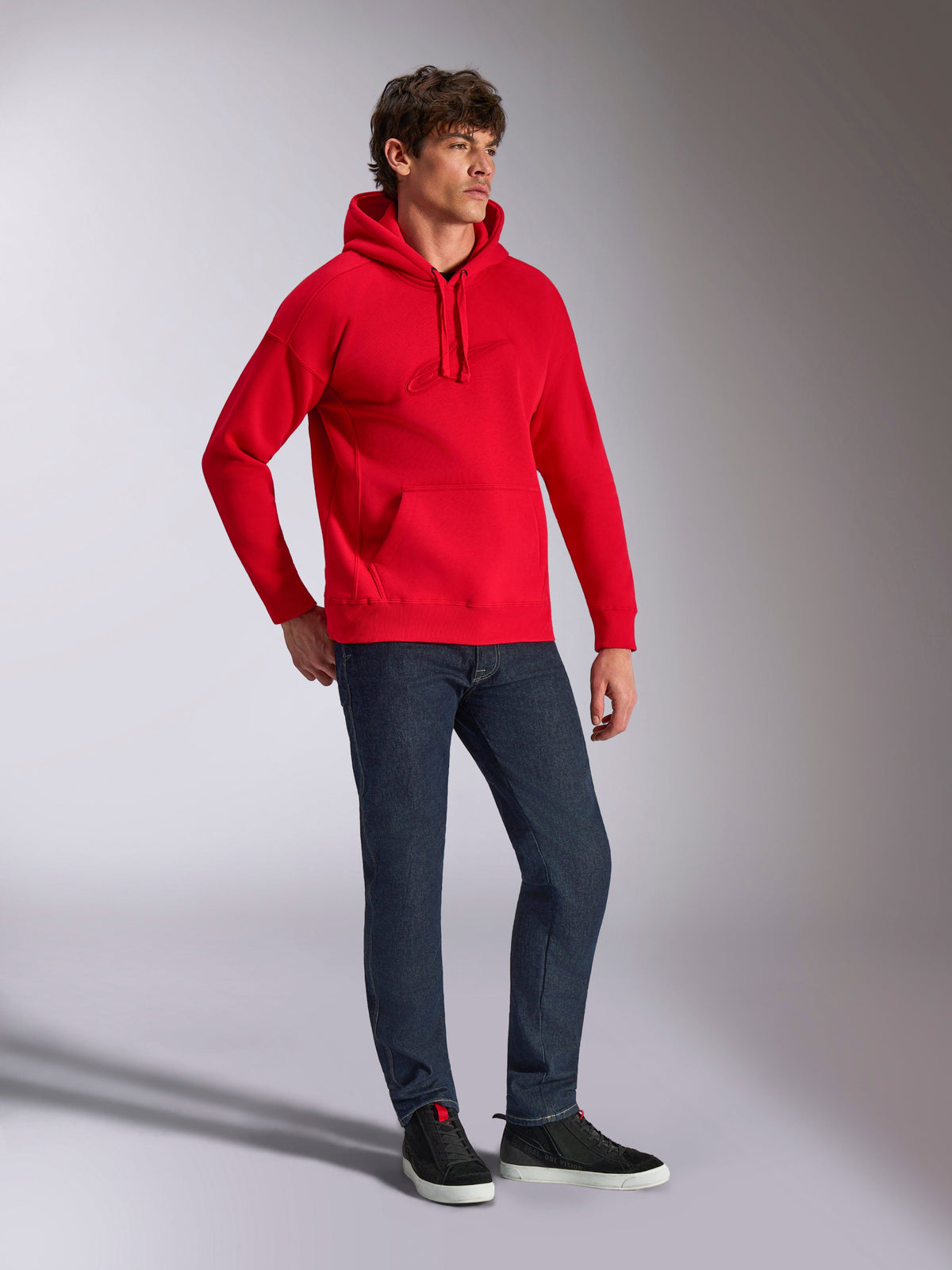 Alpinestars Emergent Oversized Hoodie, czerwona polarowa bluza z kapturem z tonalnym haftem 3D, krój oversize, noszona przez modela z ciemnoniebieskimi dżinsami i czarnymi sneakersami, codzienna odzież typu streetwear inspirowana motorsportem