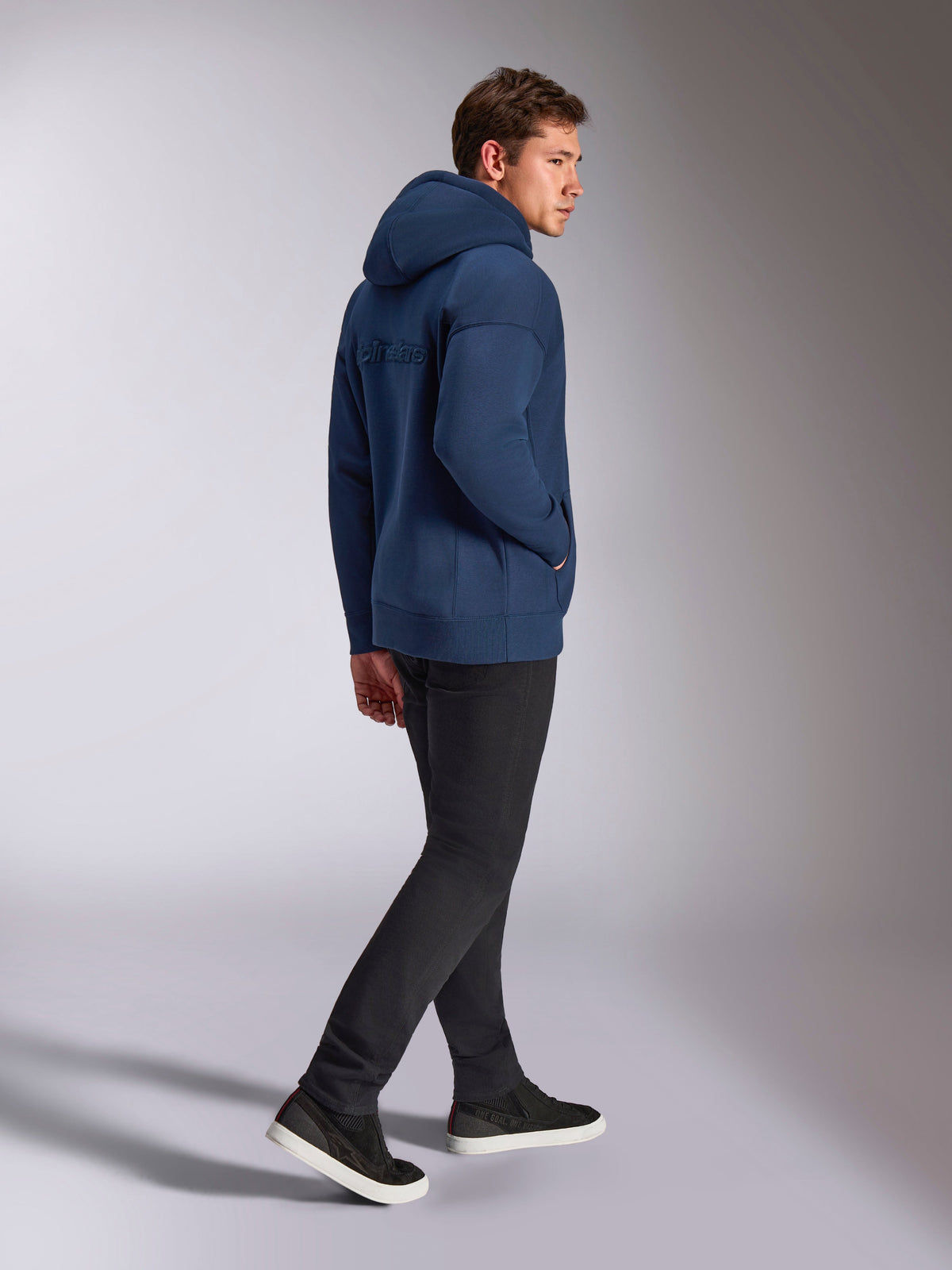 Alpinestars Emergent Oversized Hoodie, granatowa bluza z kapturem, widok z tyłu z haftowanym logo 3D Alpinestars, luźny fason z polaru, kaptur ze sznurkiem, czarne spodnie, codzienny streetwear inspirowany motorsportem