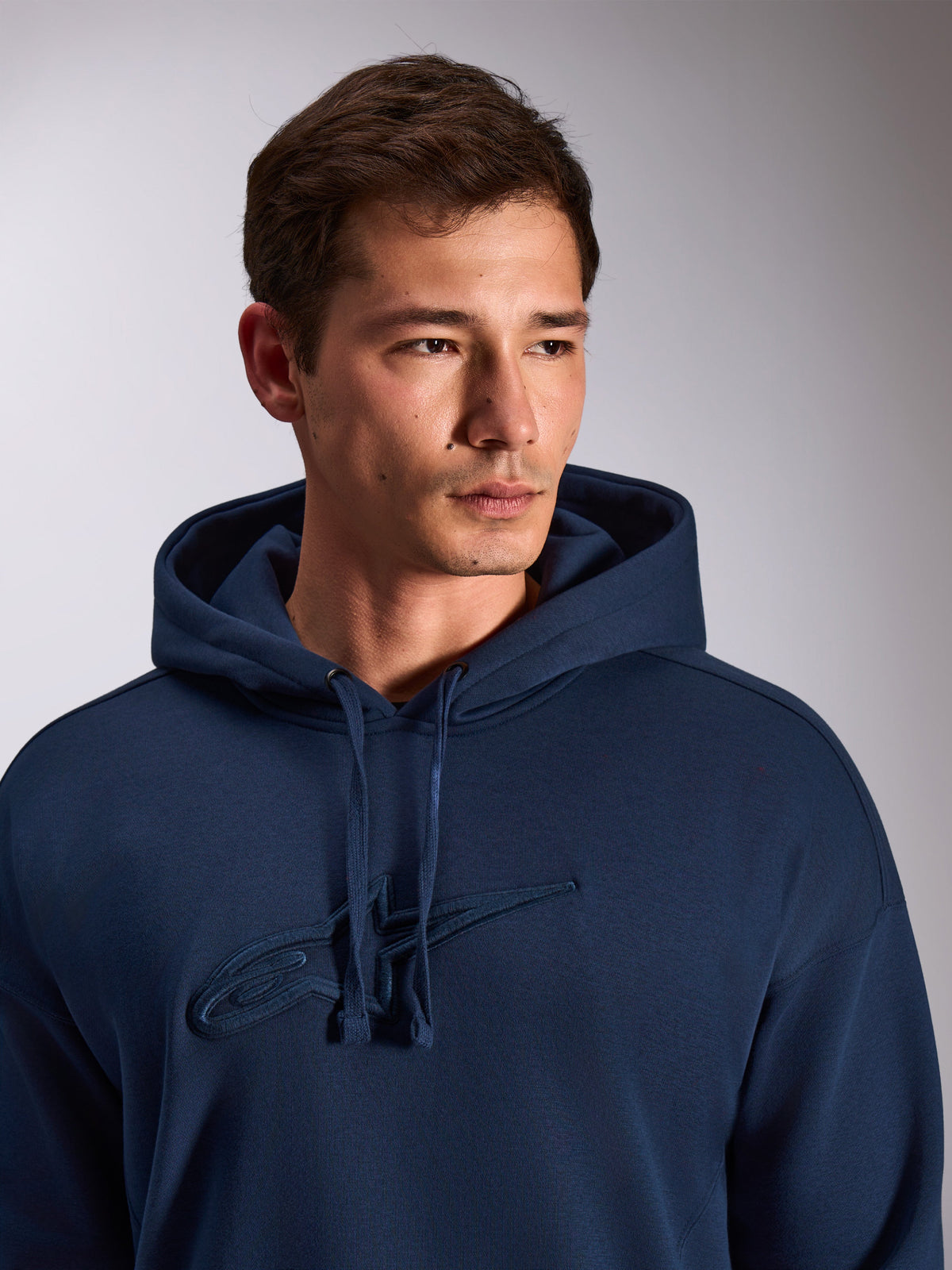 Alpinestars Emergent Oversized Hoodie, granatowa bluza z kapturem, zbliżenie na modela ubranego w polarową bluzę, tonalne haftowane logo Astars 3D na piersi, regulowany kaptur ze sznurkiem, codzienny streetwear motorsportowy