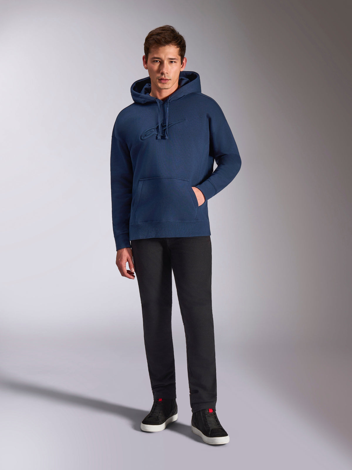Alpinestars Emergent Oversized Hoodie, granatowa bluza z kapturem z polaru, haftowane logo 3D w tym samym kolorze na przodzie, model w czarnych spodniach i sneakersach, codzienny styl streetwearowy