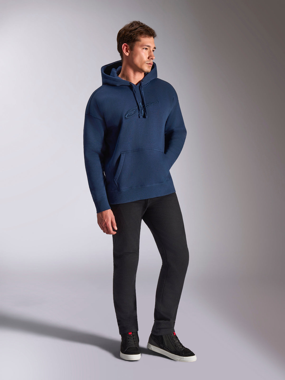 Alpinestars Emergent Oversized Hoodie, granatowa bluza z kapturem, model w czarnych spodniach i sneakersach, haftowane logo Astars 3D, kieszeń typu kangur, kaptur ze sznurkiem, casualowy streetwear inspirowany motorsportem