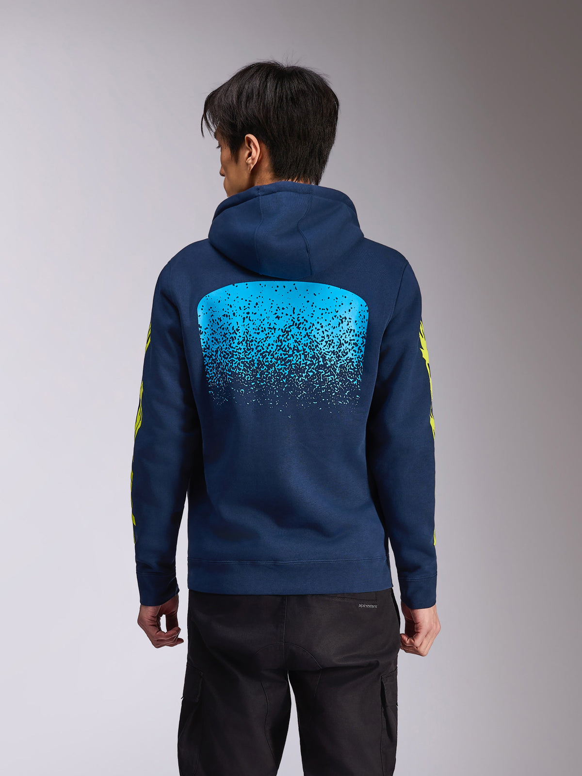 Alpinestars Obsessed Hoodie, granatowa polarowa bluza z kapturem, widok z tyłu z dużą jasnoniebieską grafiką z gradacją MX, żółty napis Alpinestars na rękawach, codzienna odzież uliczna inspirowana motorsportem
