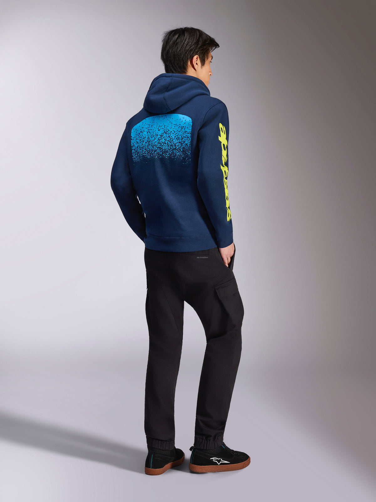 Alpinestars Obsessed Hoodie, granatowa bluza sportowa, widok z tyłu przedstawiający jasnoniebieską grafikę z gradacją MX i żółte logo Alpinestars na rękawie, noszona z czarnymi spodniami i tenisówkami