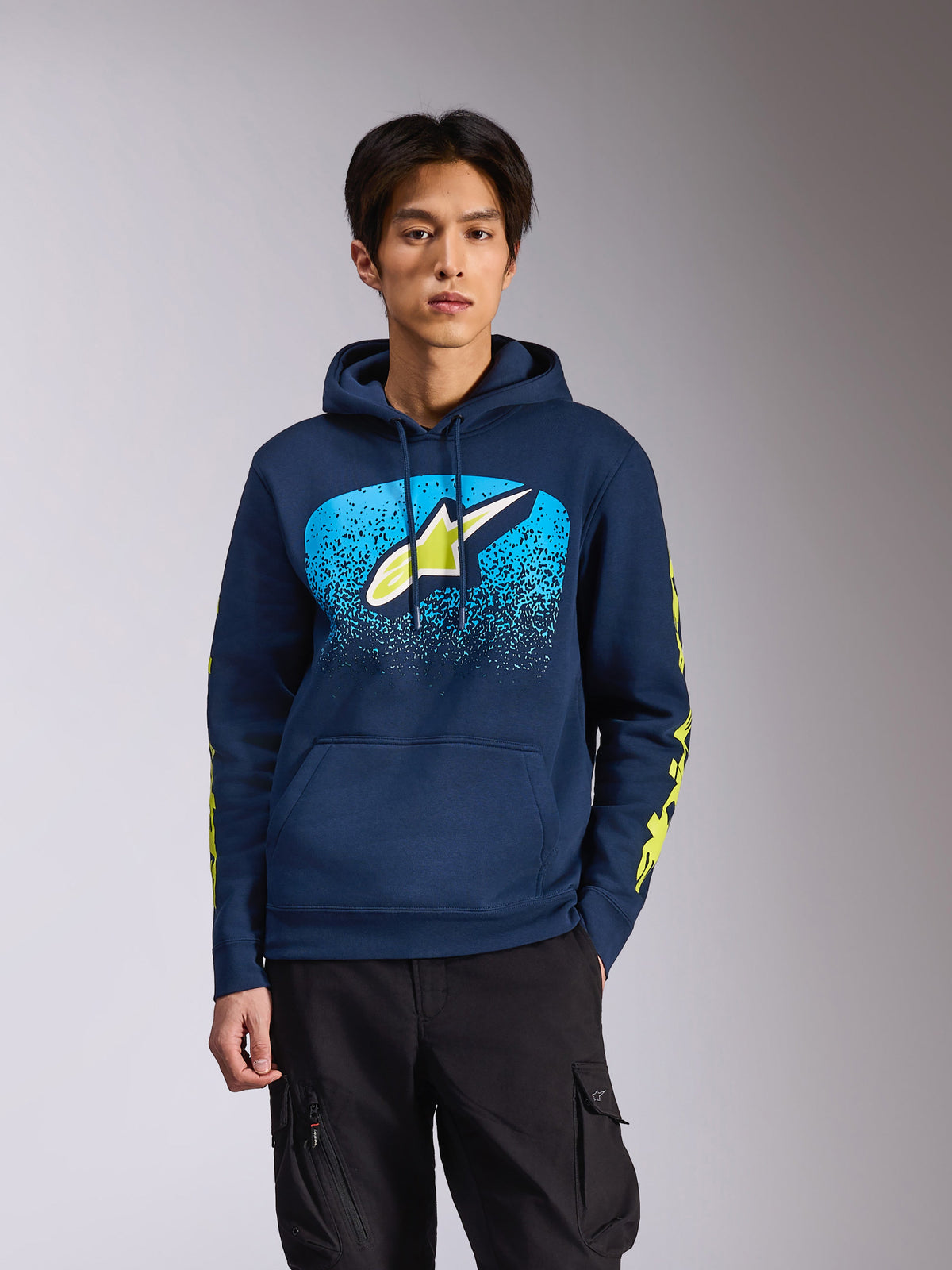 Alpinestars Obsessed Hoodie, granatowa bluza z kapturem na męskim modelu, limonkowe logo Astars z jasnoniebieską gradacją MX na piersi, żółte napisy na rękawach, casualowa odzież streetwear inspirowana motorsportem