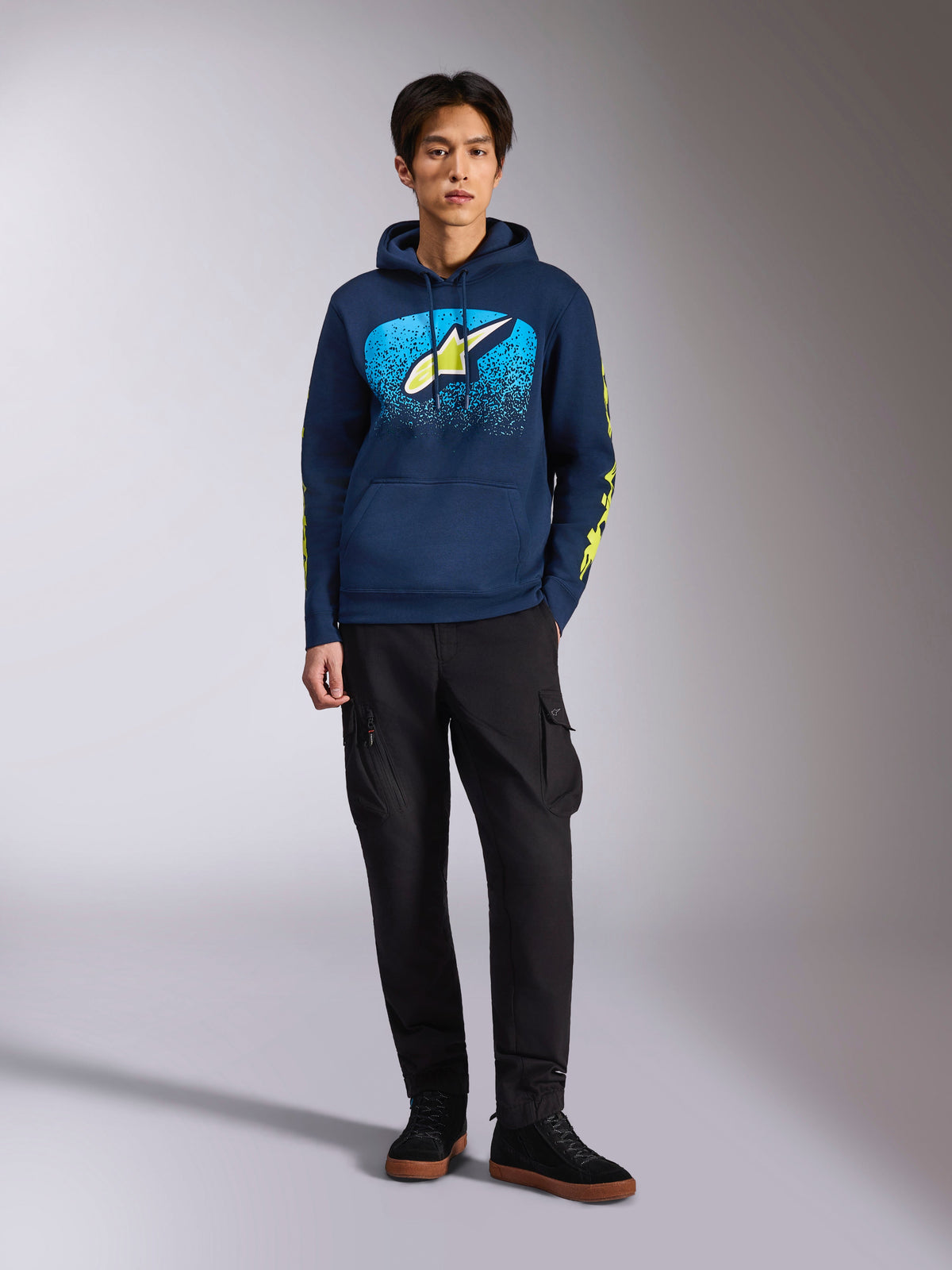 Bluza z kapturem Alpinestars Obsessed, granatowa bluza z kapturem, z przodu logo Astars z niebieską gradacją MX i limonkowo-żółtą gwiazdą, limonkowo-żółte napisy Alpinestars na rękawach, model w czarnych spodniach cargo i czarnych sneakersach, casualowy streetwear inspirowany motorsportem