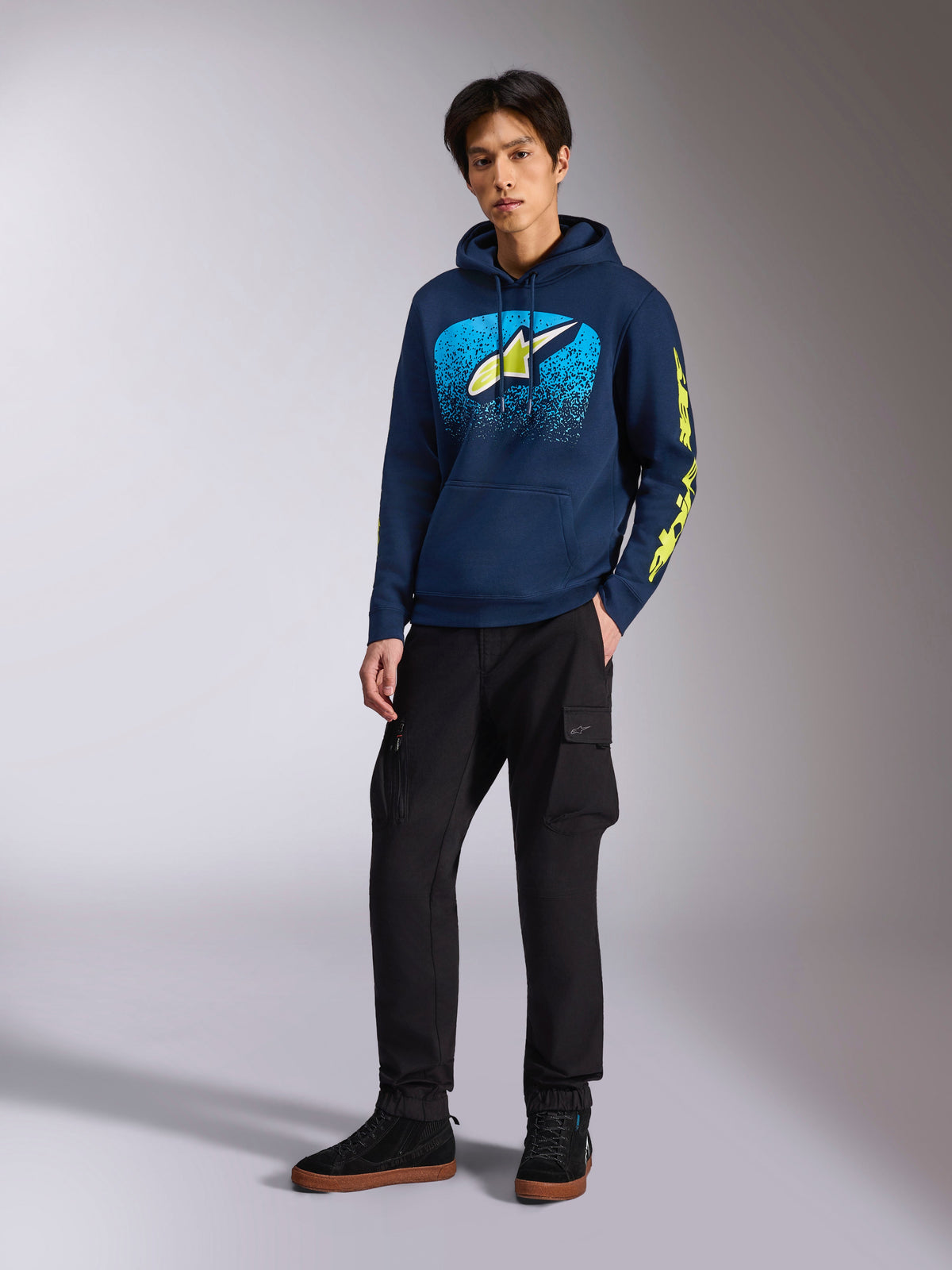 Alpinestars Obsessed Hoodie w kolorze granatowym, casualowa bluza z polaru z jasnoniebieską i limonkowo-żółtą grafiką z przodu, limonkowo-żółte logo Alpinestars na rękawie, model w czarnych spodniach cargo i czarnych sneakersach, streetwear inspirowany sportami motorowymi