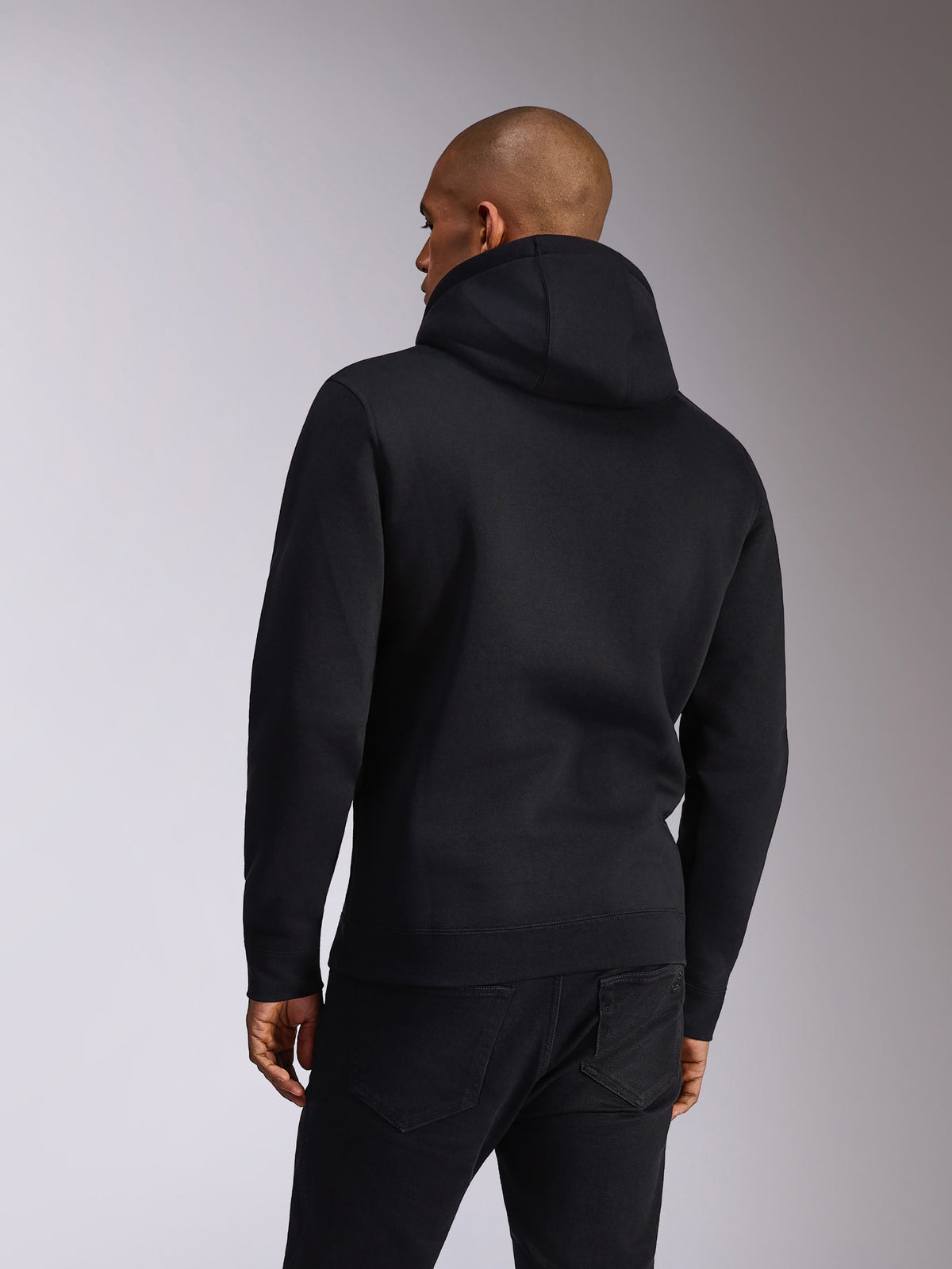 Alpinestars Ellipsoid Hoodie, czarna, widok z tyłu osoby ubranej w wkładaną przez głowę bluzę z miękkiego polaru z kapturem, długie rękawy z żebrowanymi mankietami i dołem, stylizacja z czarnymi dżinsami, codzienna odzież sportowa inspirowana sportami motorowymi
