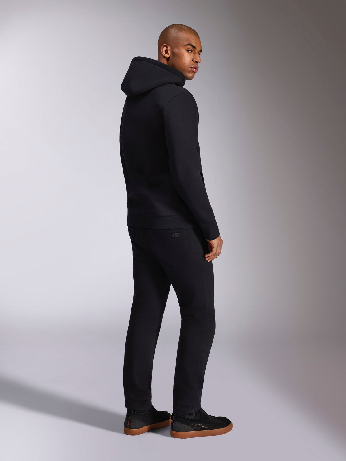 Alpinestars Ellipsoid Hoodie, czarna bluza typu streetwear, widok z tyłu na modelu, polarowa bluza z kapturem i ściągaczami, zestawiona z czarnymi spodniami i sneakersami, nowoczesny design inspirowany motorsportem