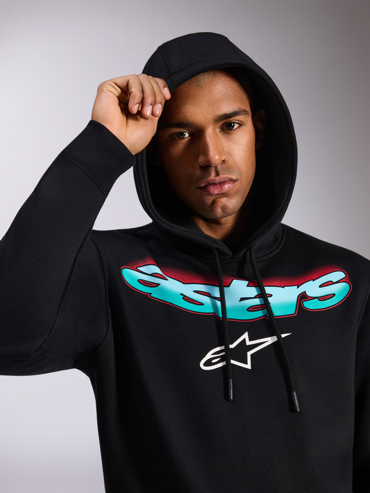 Alpinestars Ellipsoid Hoodie, czarna bluza z polaru z kapturem, stylizowany turkusowo-czerwony napis Astars i białe logo na piersi, noszona przez modela z założonym kapturem, codzienna odzież uliczna inspirowana sportami motorowymi