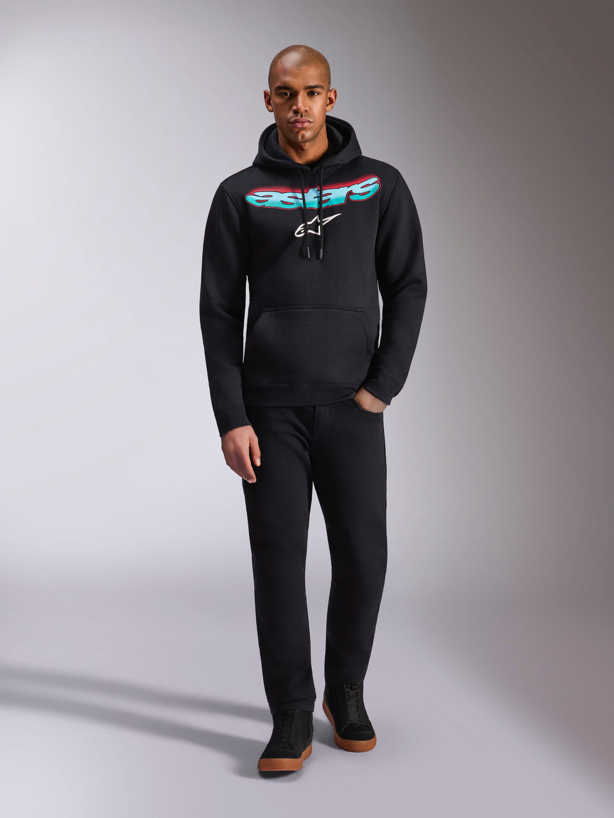 Alpinestars Ellipsoid Hoodie, czarna bluza z kapturem z polaru, zakrzywiony napis Astars z czerwono-turkusowym gradientem i białym logo gwiazdy, model w czarnych spodniach i sneakersach, codzienna odzież streetwearowa w stylu motorsportowym
