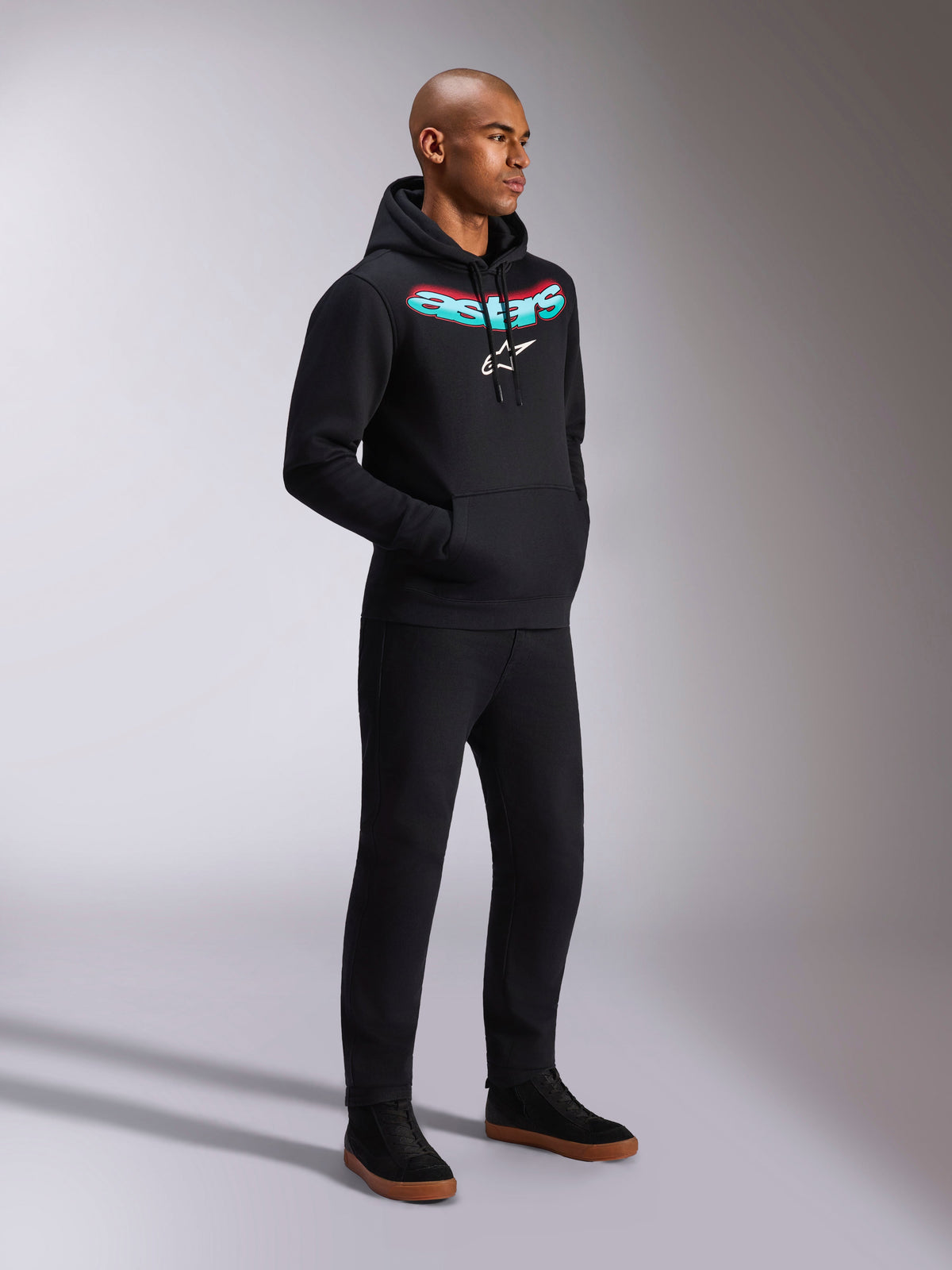 Alpinestars Ellipsoid Hoodie, czarna bluza z kapturem z polaru, z zakrzywionym napisem Astars z morsko-czerwonym gradientem i białym logo gwiazdy, noszona przez modela do czarnych spodni, codzienna odzież uliczna inspirowana sportami motorowymi
