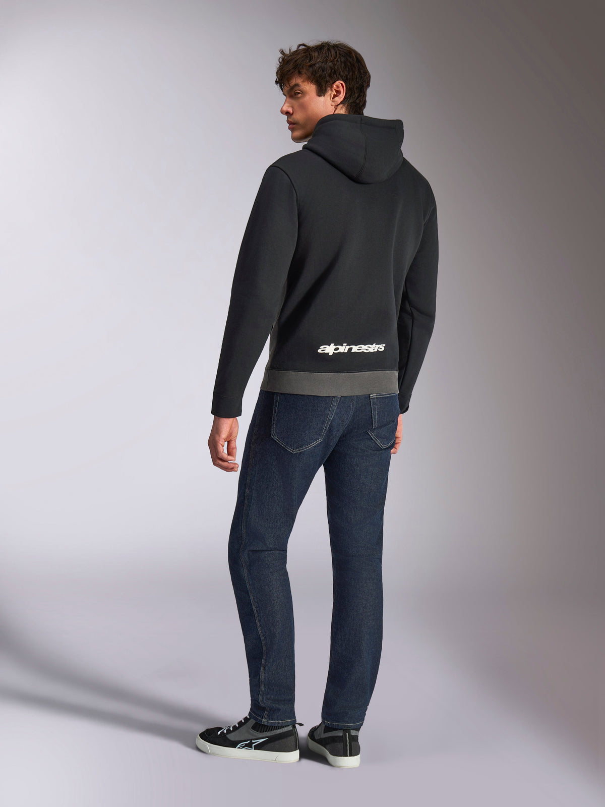 Alpinestars Parallel Hoodie, rear view of model wearing the black and gunmetal gray pullover, featuring white Alpinestars logo on the lower back, styled with dark blue jeans, casual motorsport-inspired streetwear