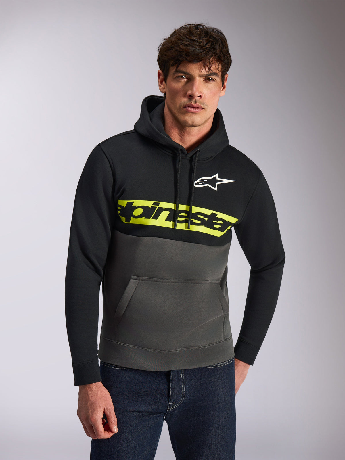 Alpinestars Parallel Hoodie, casual streetwear, color-blocked black and gunmetal grey, bright yellow chest stripe with Alpinestars word logo, white Astars logo, worn by a model with blue jeans, modern motorsport-inspired design
