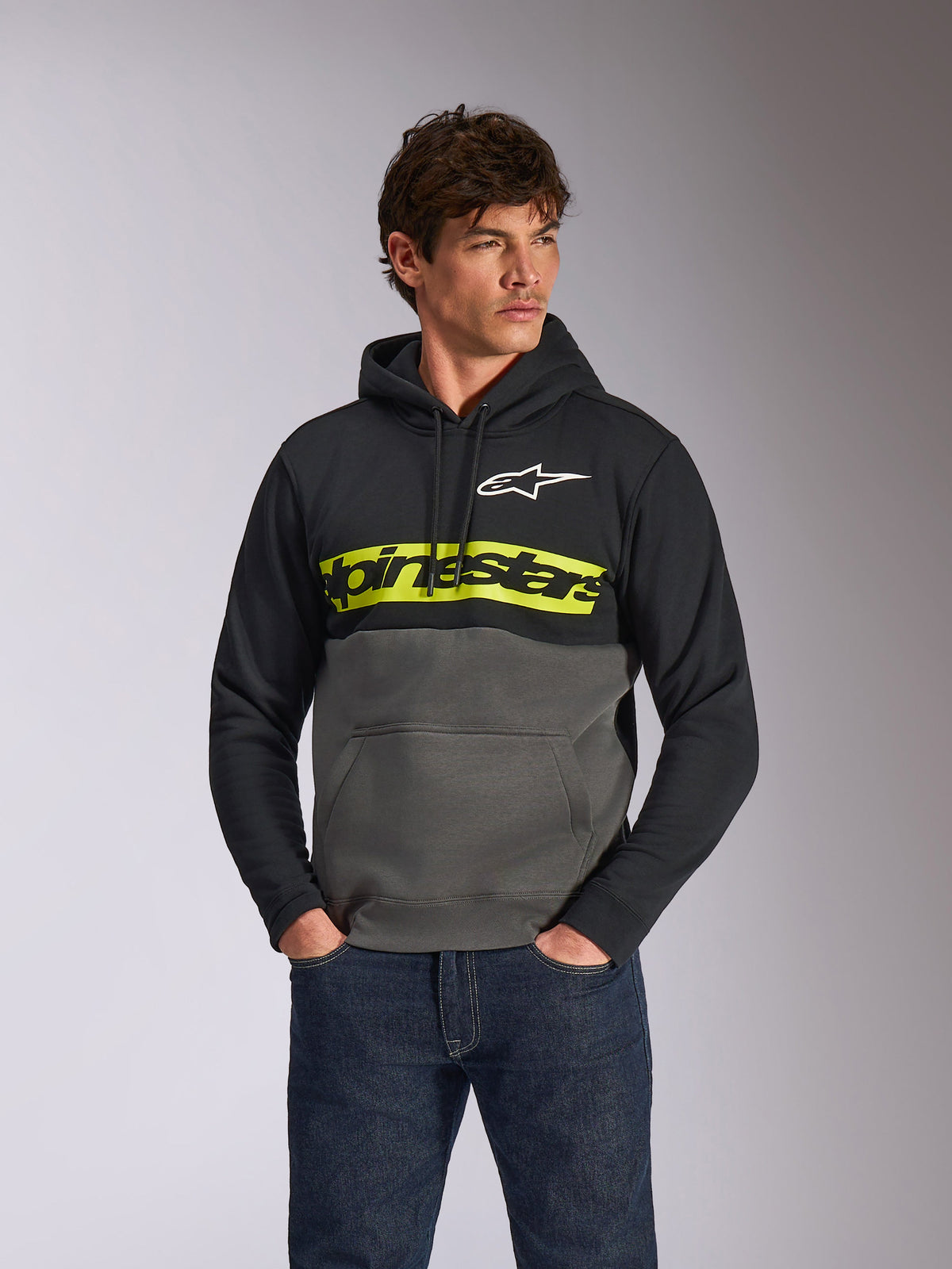 Alpinestars Parallel Hoodie, casual color-blocked pullover fleece, Black and Gunmetal with a vibrant yellow chest bar, Alpinestars word logo and white star logo, worn by a male model with dark blue jeans