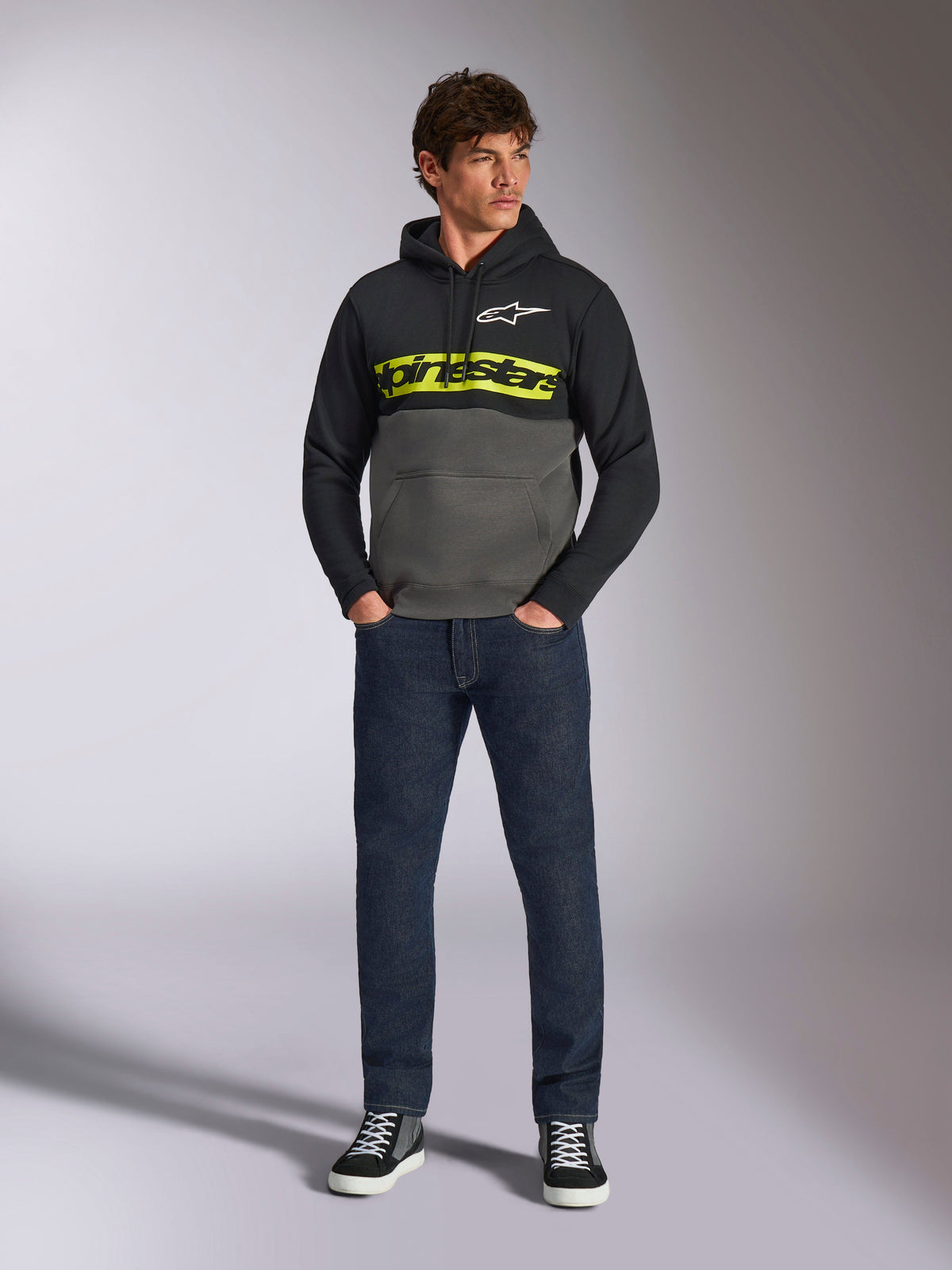 Alpinestars Parallel Hoodie, color-blocked pullover, Black and Gunmetal gray, bright yellow Alpinestars word logo and white star logo on chest, worn by a male model with dark blue jeans and black sneakers, casual motorsport streetwear