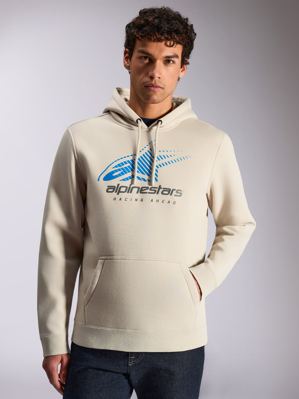 Bluza z kapturem Alpinestars Pulse, codzienny streetwear, kolor piaskowy, z przodu niebiesko-szare logo Astars Combo z efektem zanikających linii i napisem „Racing Ahead”, kaptur ze sznurkiem, kieszeń typu kangurka, noszona przez modela w ciemnych jeansach