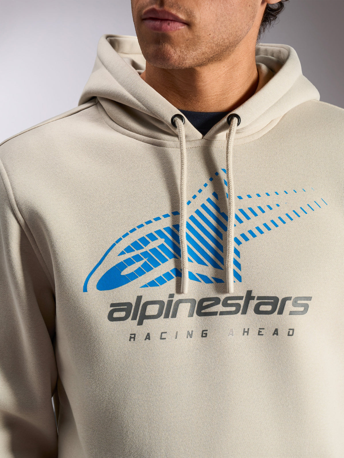 Alpinestars Pulse Hoodie, codzienna bluza typu pullover w kolorze złamanej bieli, zbliżenie na klatkę piersiową z niebiesko-szarym logo Astars Combo z efektem linii prędkości i napisem „Racing Ahead”, kaptur ze sznurkami z powlekanymi końcówkami