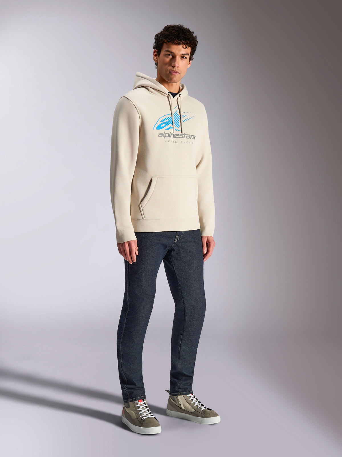 Alpinestars Pulse Hoodie, codzienna bluza streetwearowa w kolorze Off White, polarowy fason z kapturem, niebiesko-szare logo Astars z efektem przejścia i liniami prędkości, kieszeń typu kangurka, model w ciemnych jeansach i wysokich butach sportowych