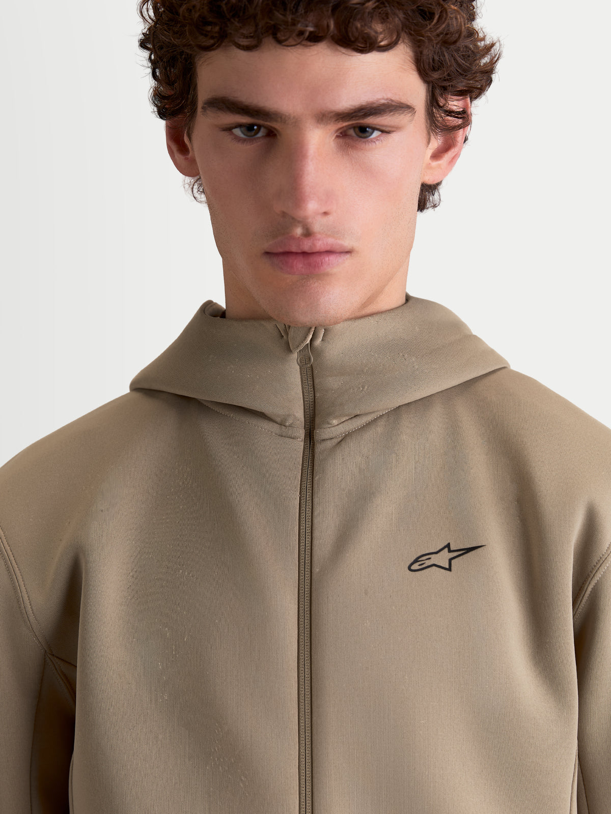Alpinestars bluza z kapturem Cypher, beżowa sportowa bluza z wysokim kapturem prezentowana na modelu, pełny zamek, małe czarne logo Alpinestars na piersi, techniczna tkanina tri-blend, codzienna odzież sportowa