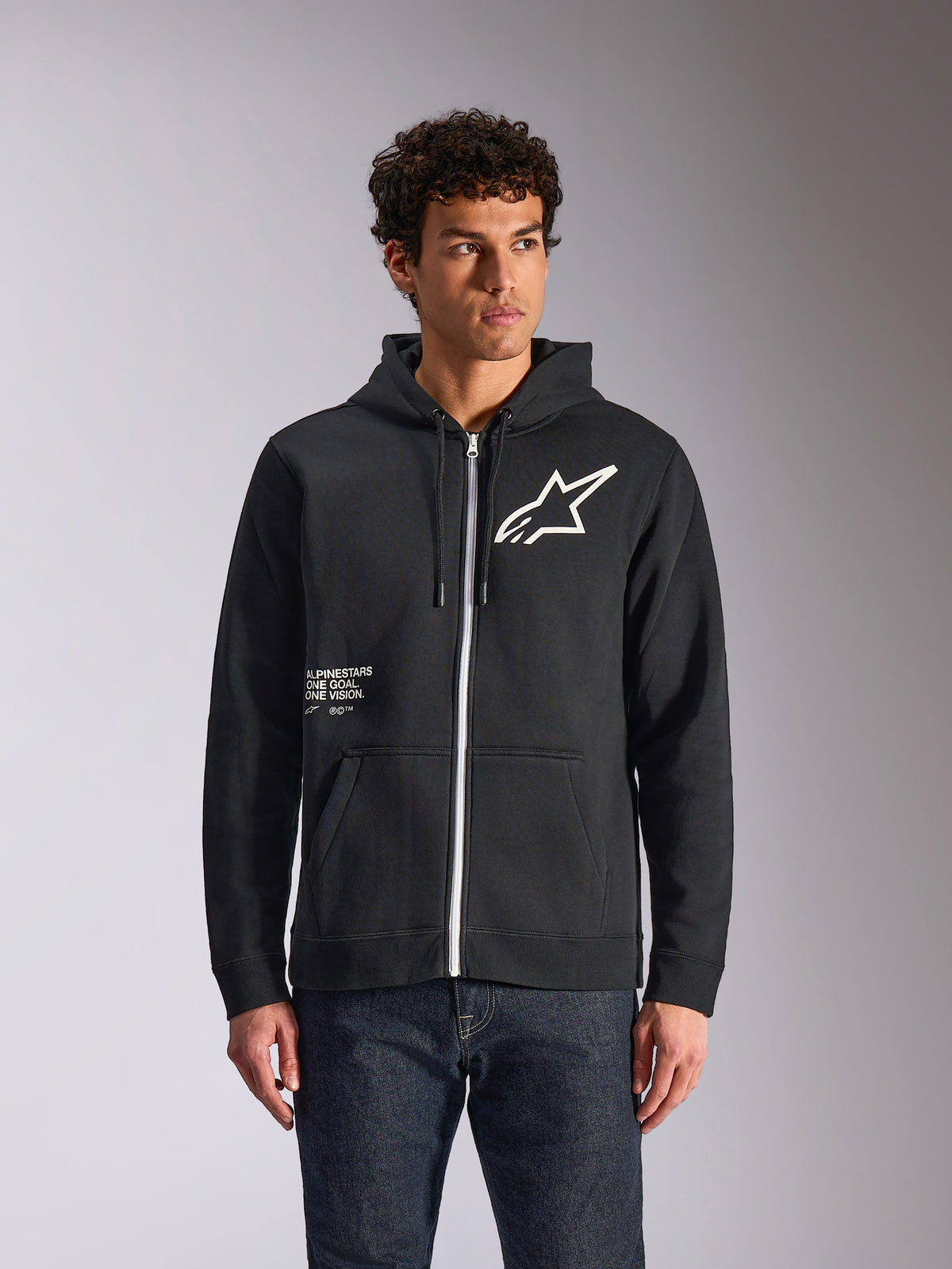 Alpinestars Transverse Hoodie, czarna bluza z kapturem zapinana na zamek, białe logo Astars i grafika One Goal z przodu, model w ciemnych dżinsach, codzienna odzież uliczna inspirowana sportami motorowymi