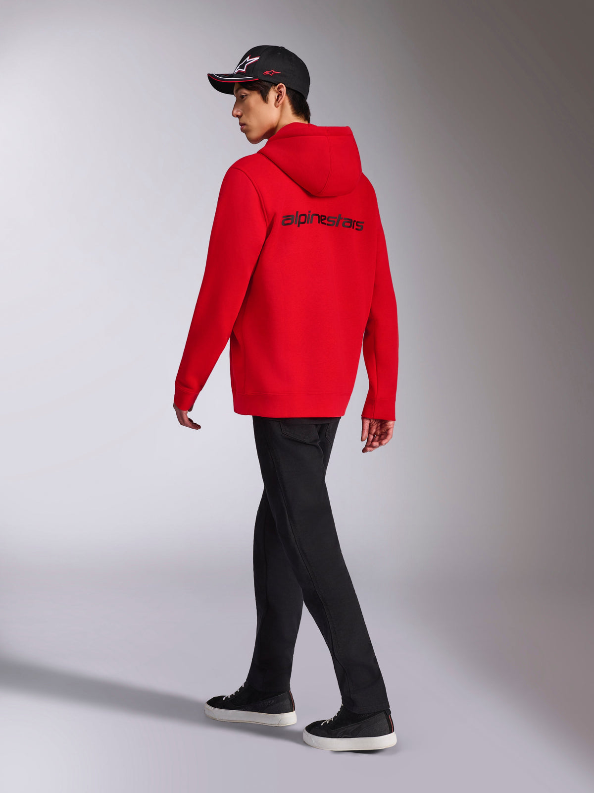 Alpinestars Transverse Hoodie, czerwona rozpinana bluza z polaru z kapturem, widok z tyłu z czarnym logotypem Alpinestars, noszona przez modela w czarnych spodniach i czarnej czapce z logo, casualowy streetwear inspirowany motorsportem