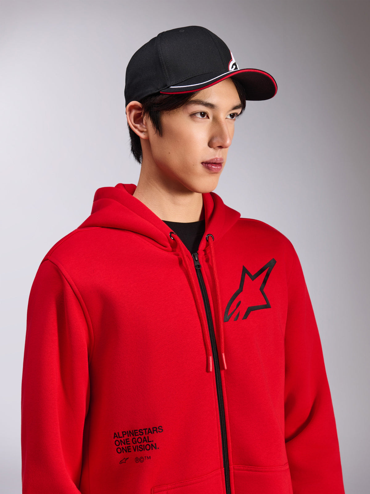 Bluza z kapturem Alpinestars Transverse, czerwony polar zapinany na zamek, czarne logo Astars na piersi i napis „One Goal. One Vision.” na dole z przodu, casualowa odzież streetwearowa, model w czarnej czapce z daszkiem