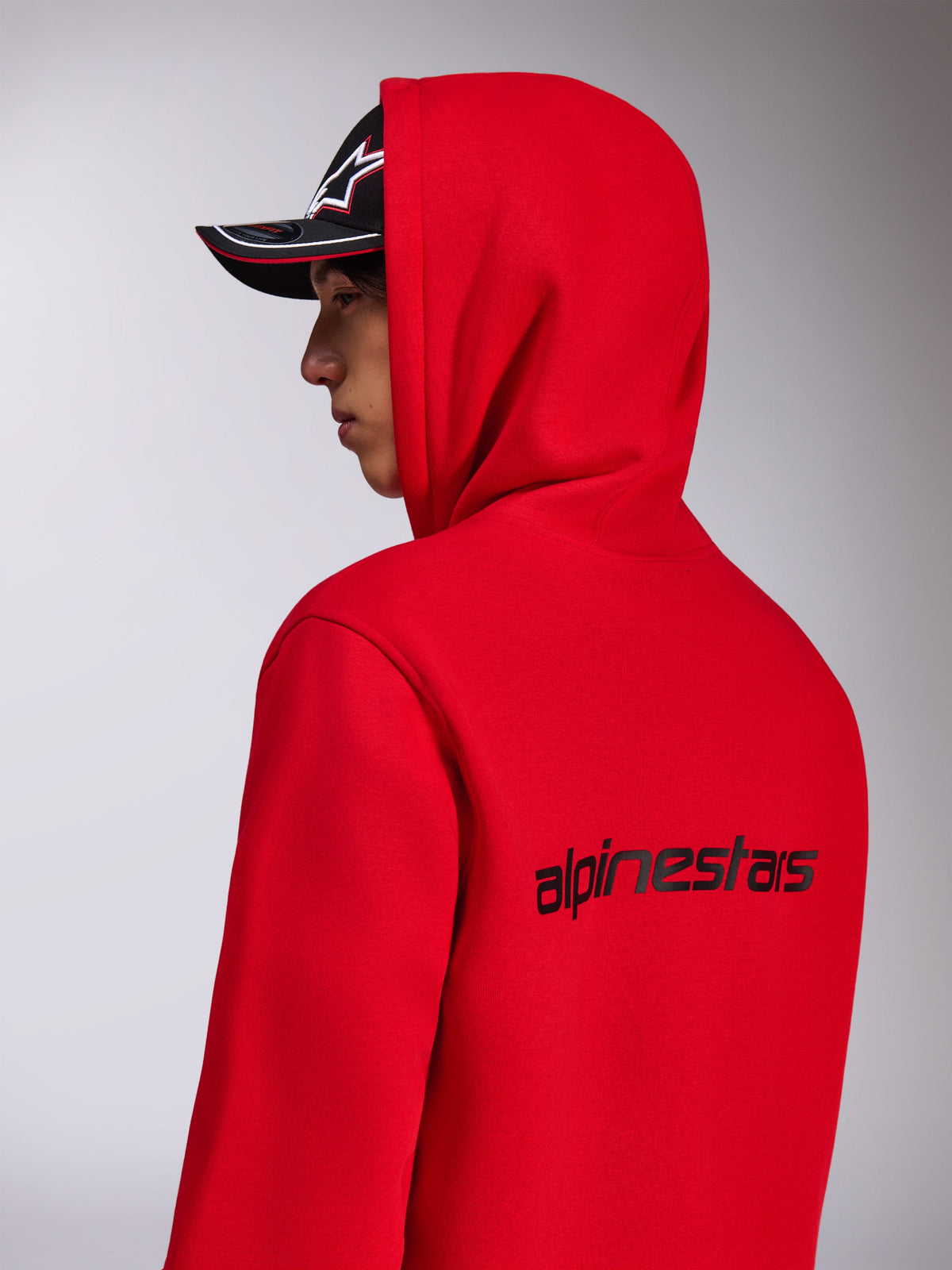Alpinestars Transverse Hoodie, czerwona bluza z kapturem z polaru, widok z boku i z tyłu ukazujący czarny napis Alpinestars na plecach, model z założonym kapturem i czarną czapką z daszkiem, codzienna odzież uliczna inspirowana sportami motorowymi
