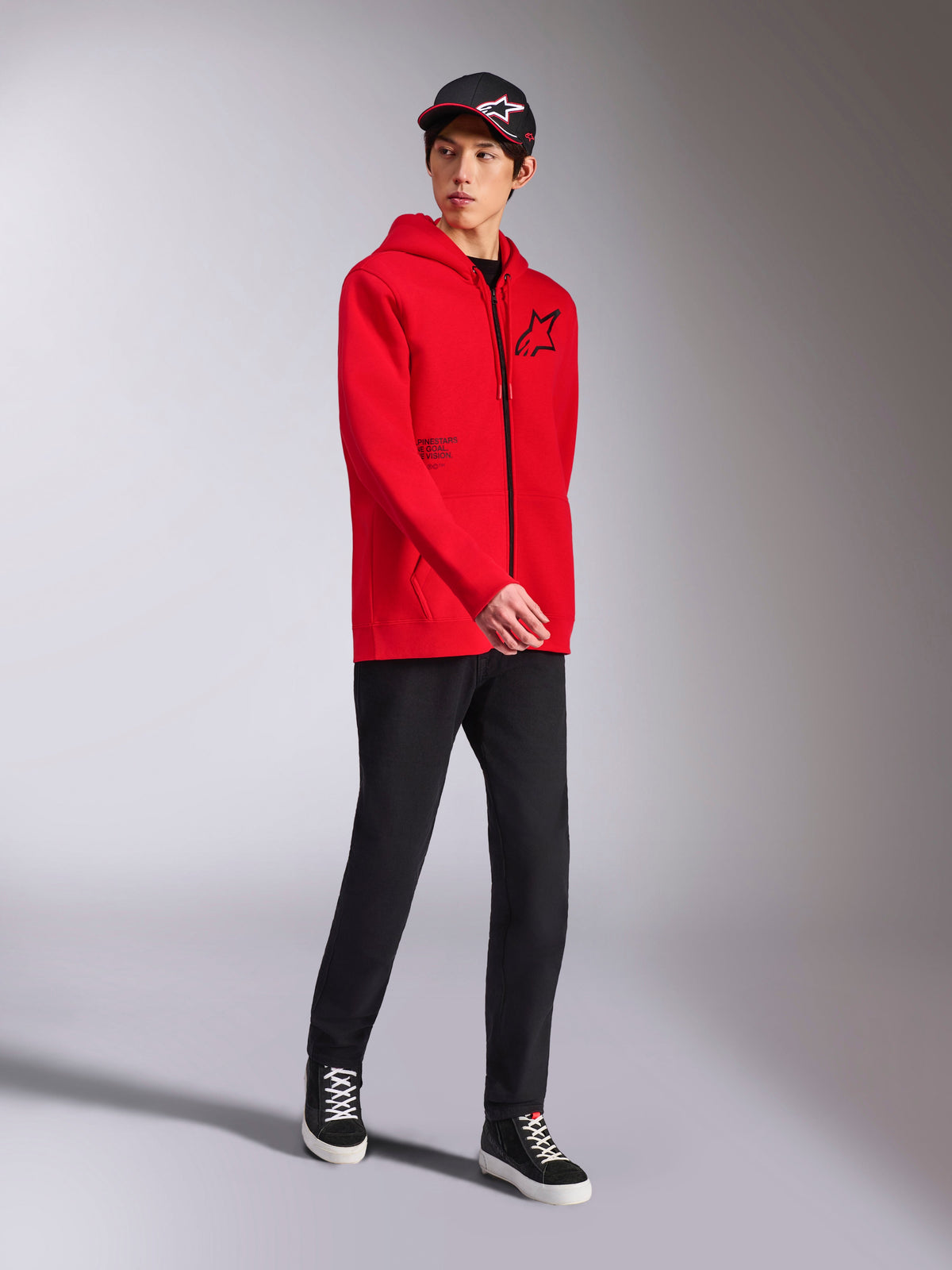 Alpinestars Transverse Hoodie w kolorze czerwonym, męski model w bluzie z kapturem z polaru zapinanej na zamek, czarne logo Astars i nadruk tekstowy, stylizacja z czarnymi spodniami i czapką z daszkiem, codzienna odzież uliczna w stylu motorsport